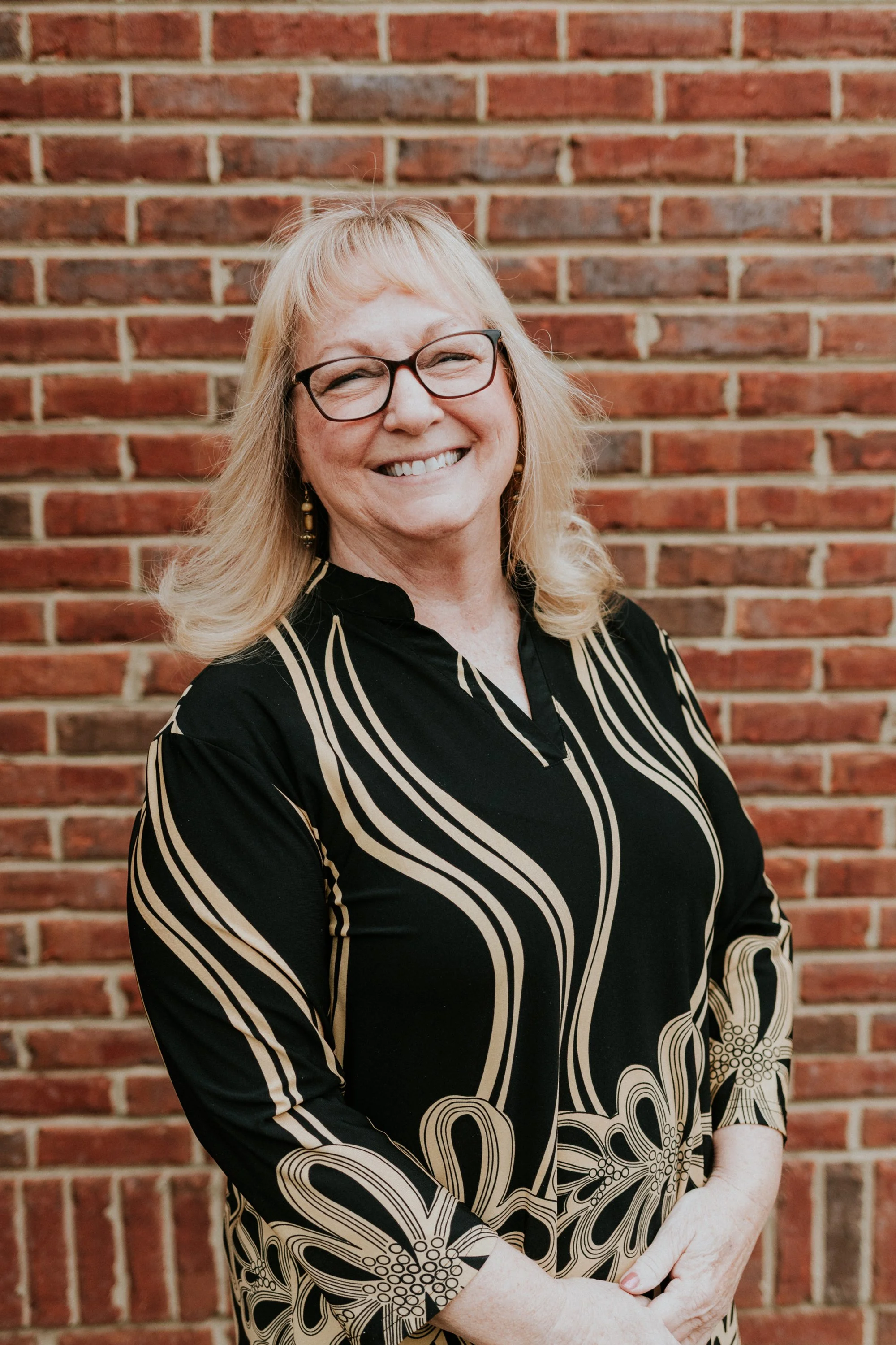 Smiling woman with glasses and blonde hair standing in front of a brick wall, wearing a black dress with beige floral and stripe pattern.