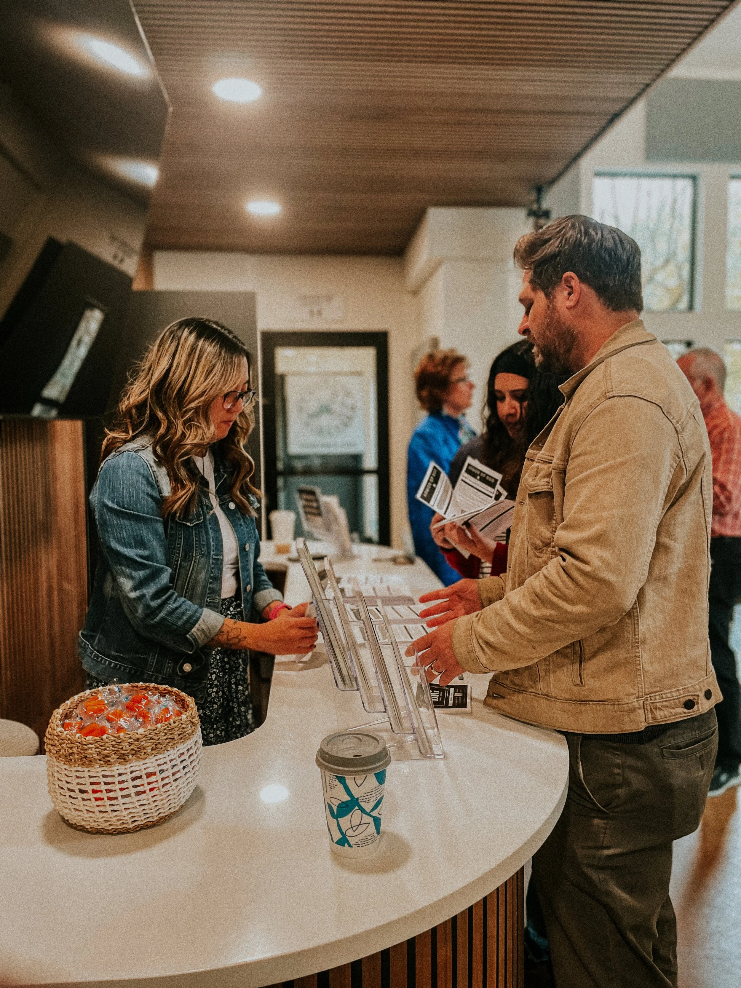 People checking in at a reception desk in a modern building, with a basket of candy and a coffee cup on the counter.