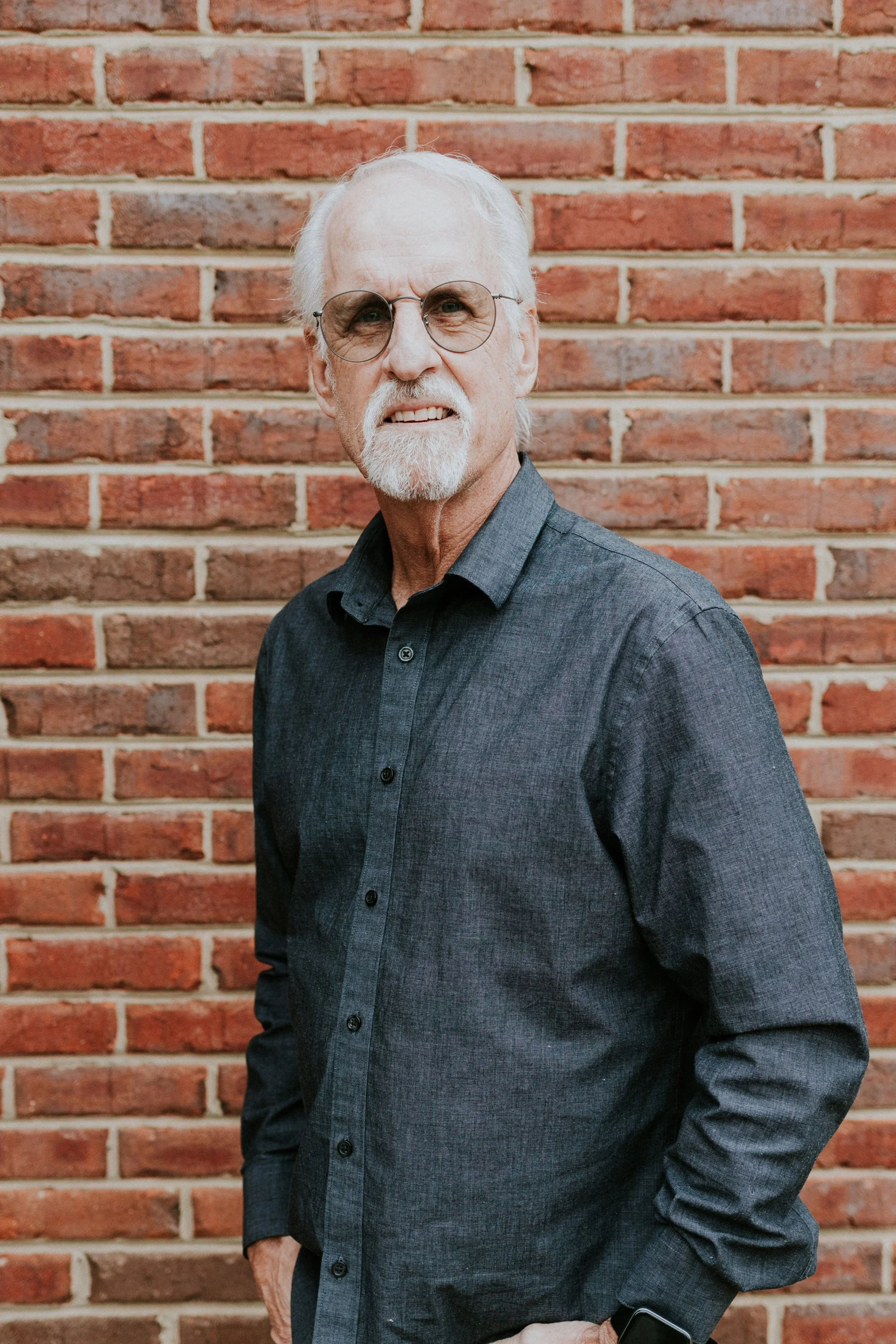 An older man with white hair, glasses, and a beard, wearing a dark shirt, standing against a brick wall.