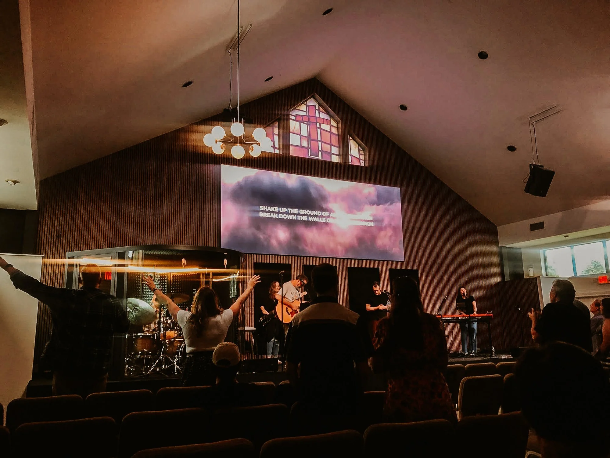 People worshipping and singing in a church during a service, with a band playing on stage, a screen displaying lyrics, and stained glass windows in the background.
