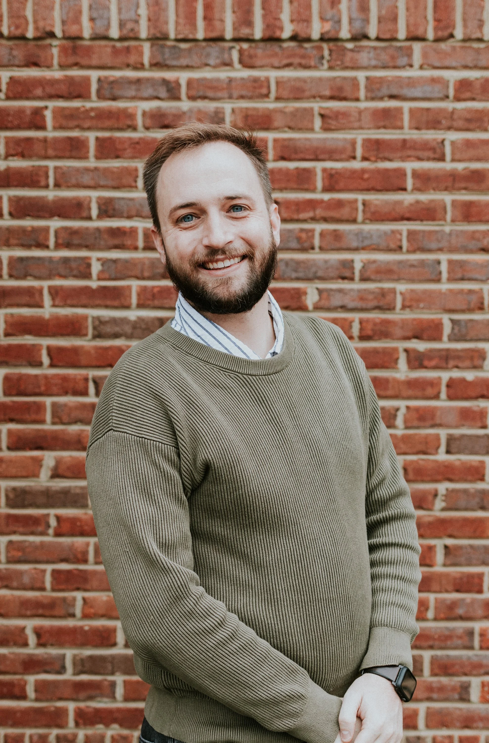A smiling man with a beard and blue eyes standing in front of a red brick wall, wearing a green sweater over a collared shirt, with a smartwatch on his left wrist.