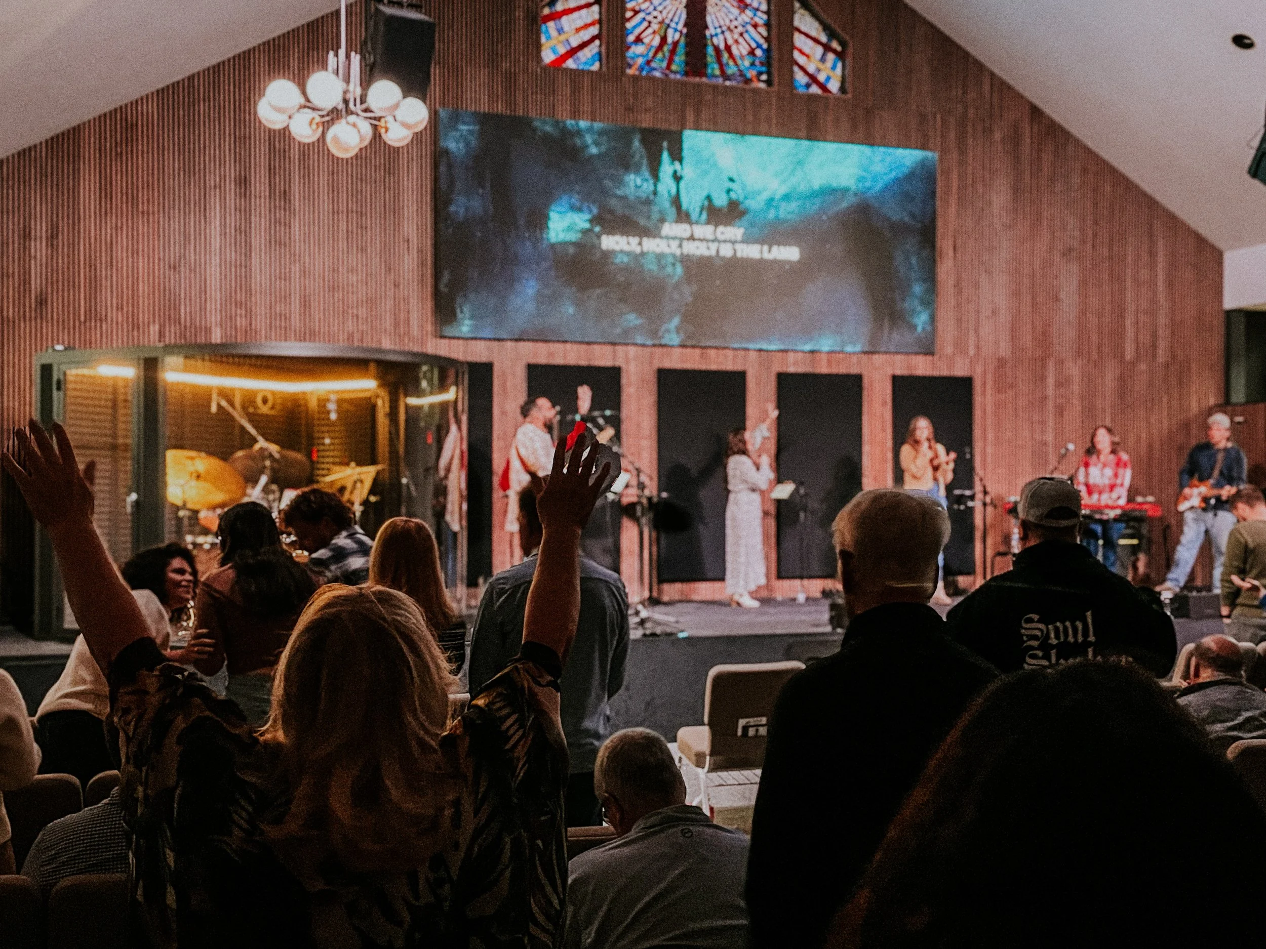 People at a church or concert hall watching a performance on stage with a band and a singer, some raising their hands, with a large screen and stained glass window above.