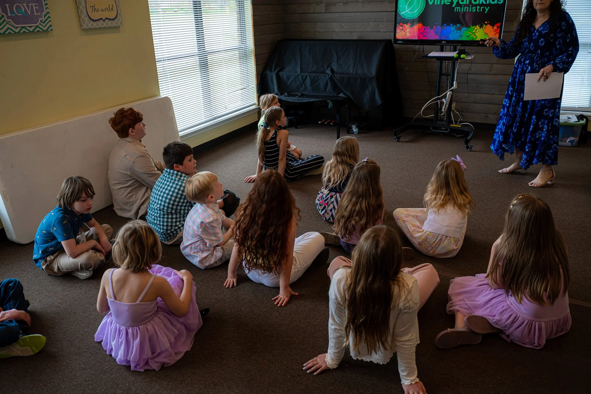 Children sitting on the carpeted floor watching a woman presenting using a TV screen that displays the logo of Vineyard Kids ministry. The woman is standing next to the TV holding a paper, and the room has large windows with blinds.