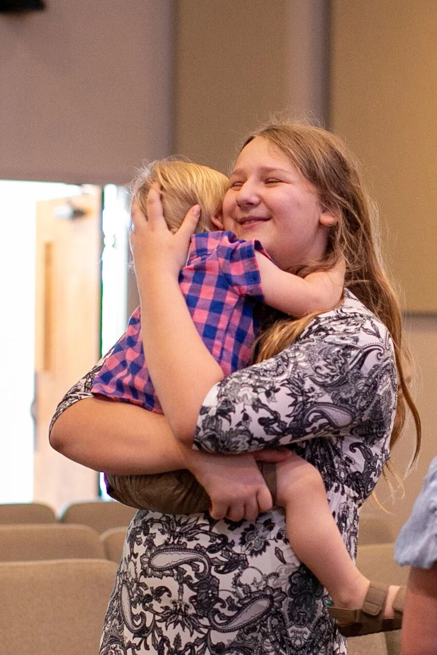 A woman with long hair, wearing a patterned dress, happily hugging a young child with blonde hair, inside a room.