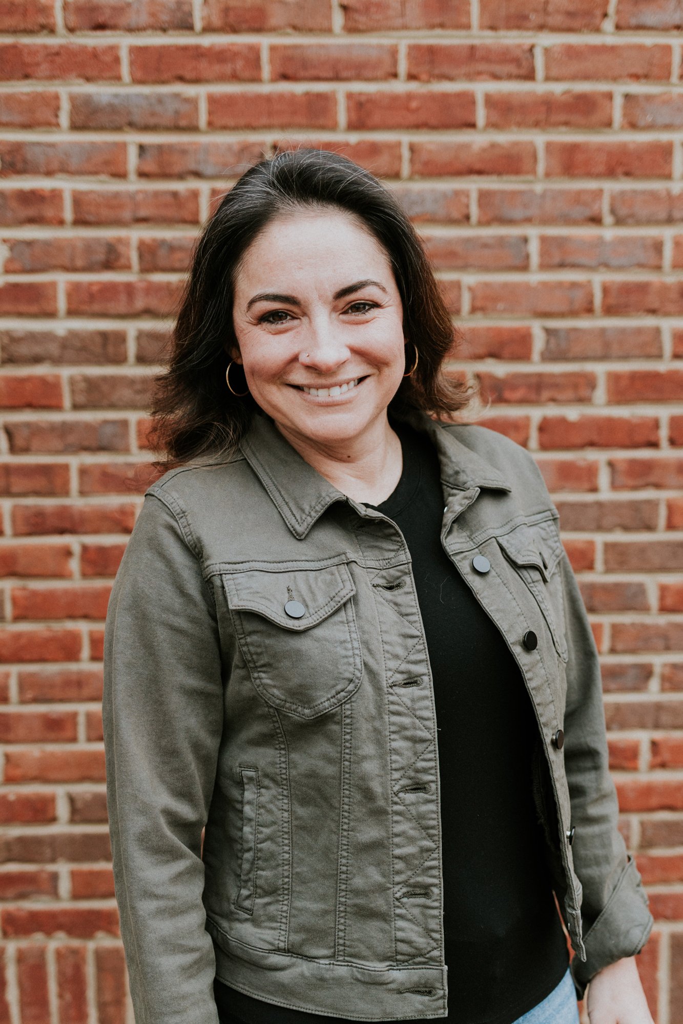 A woman with short dark hair smiling in front of a red brick wall. She is wearing an olive green denim jacket over a light gray T-shirt.