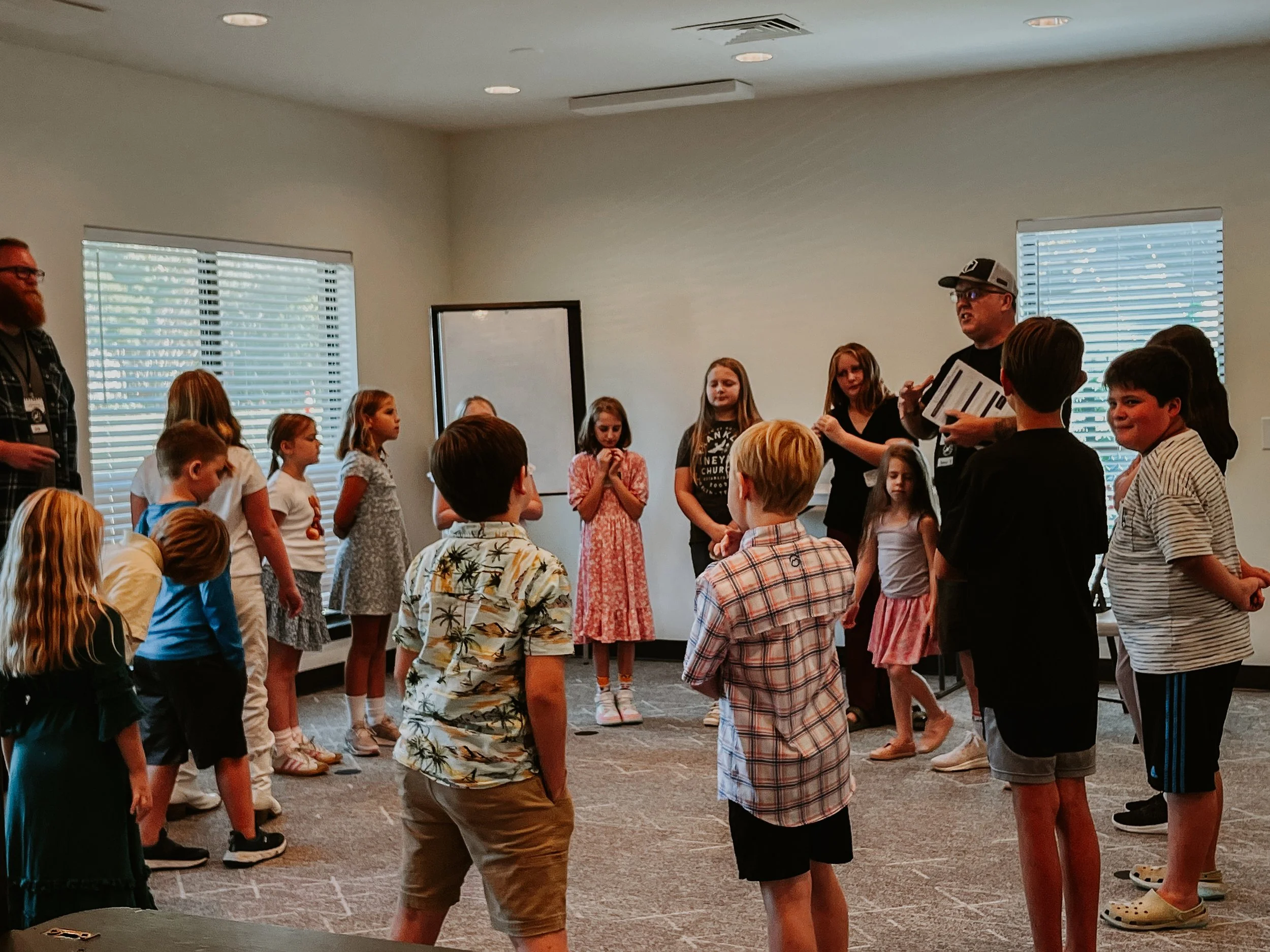 Children standing in a semi circle in a room, listening to an adult speaker who is holding a clipboard, with another adult on the side near windows with blinds.