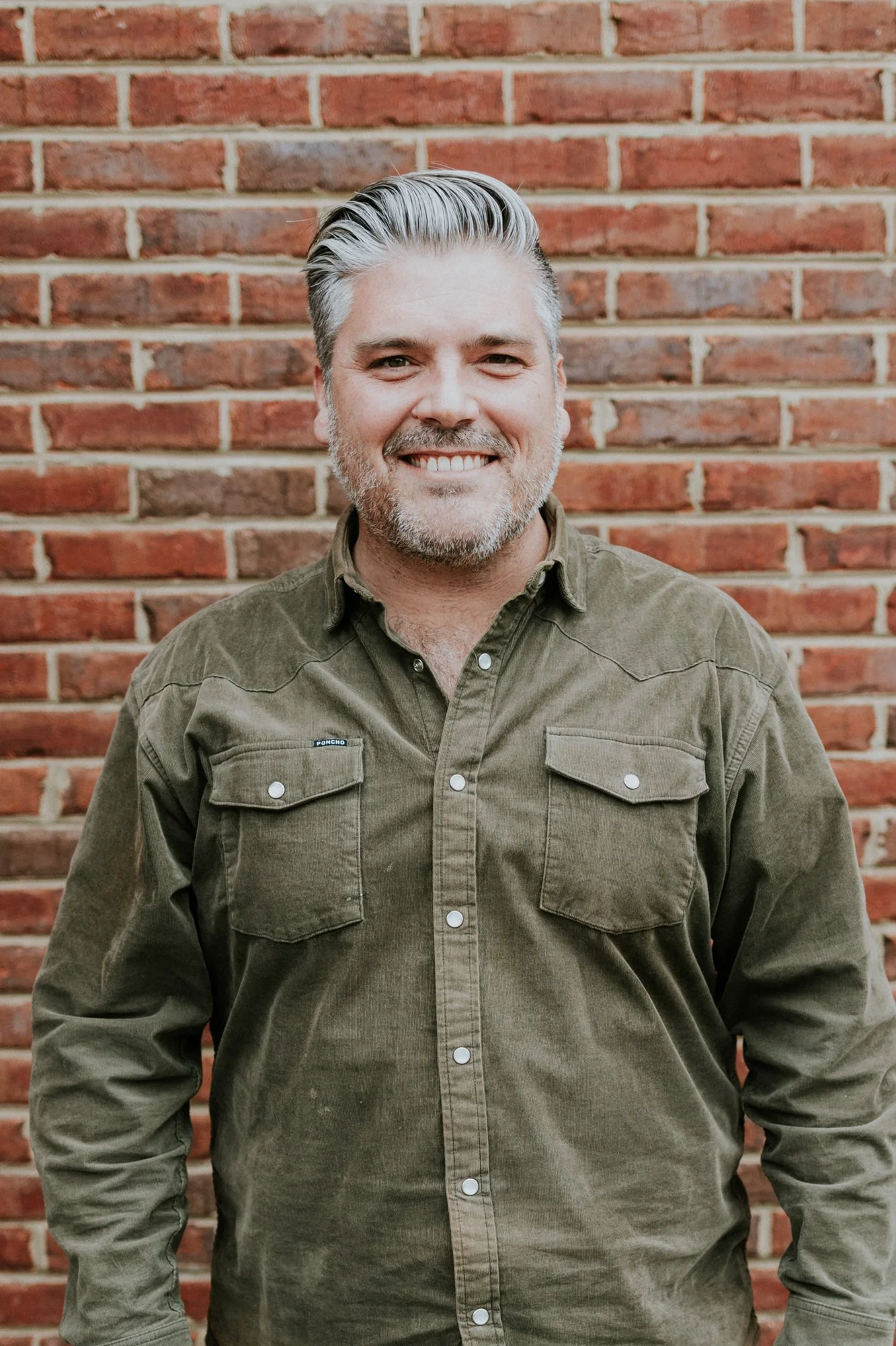 A man with salt and pepper hair and beard smiling, wearing a black shirt and a black and blue checkered shirt, standing in front of a red brick wall.