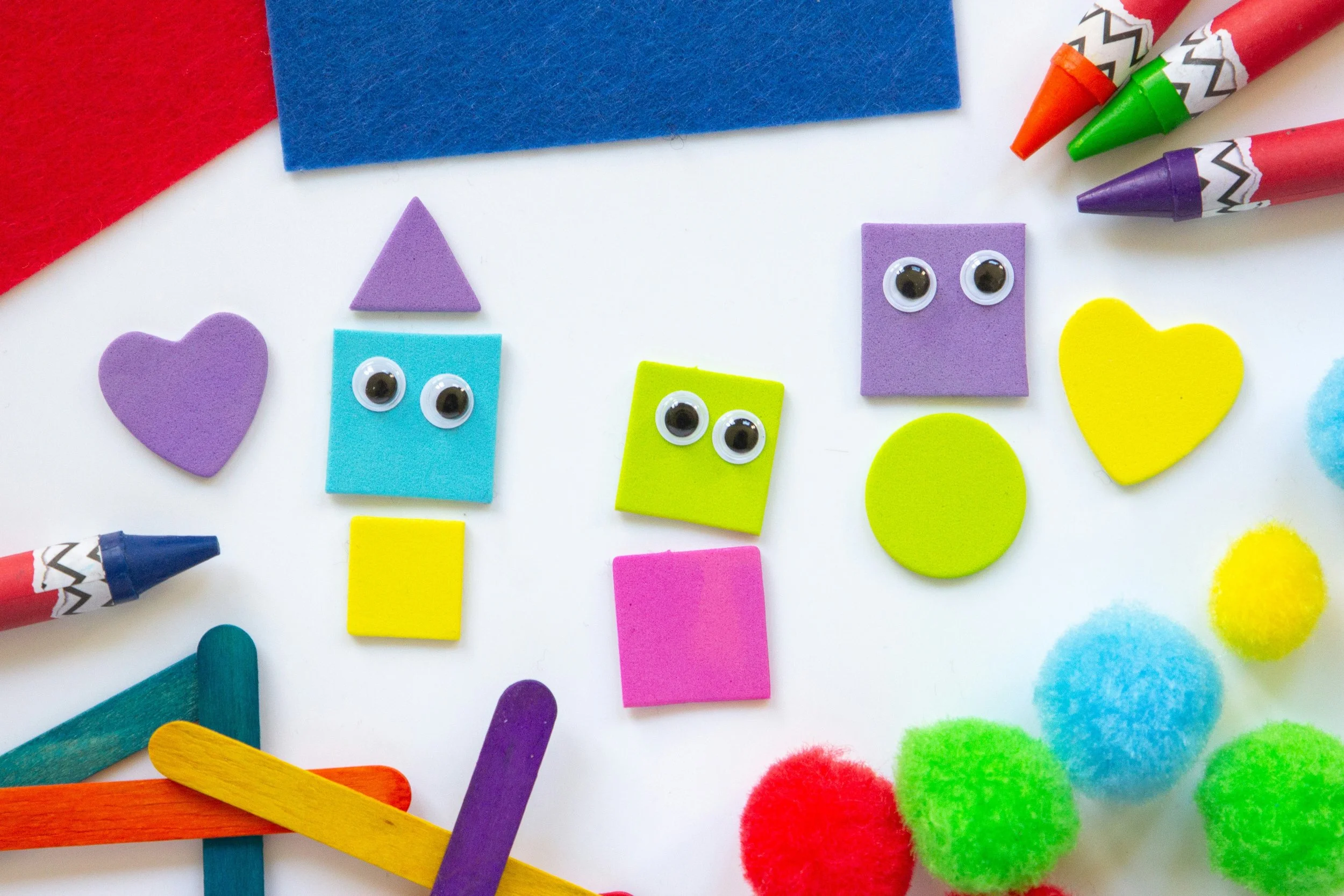 Colorful foam shapes and craft supplies arranged on a white surface, including googly eyes, felt shapes, pom-poms, and colored sticks.
