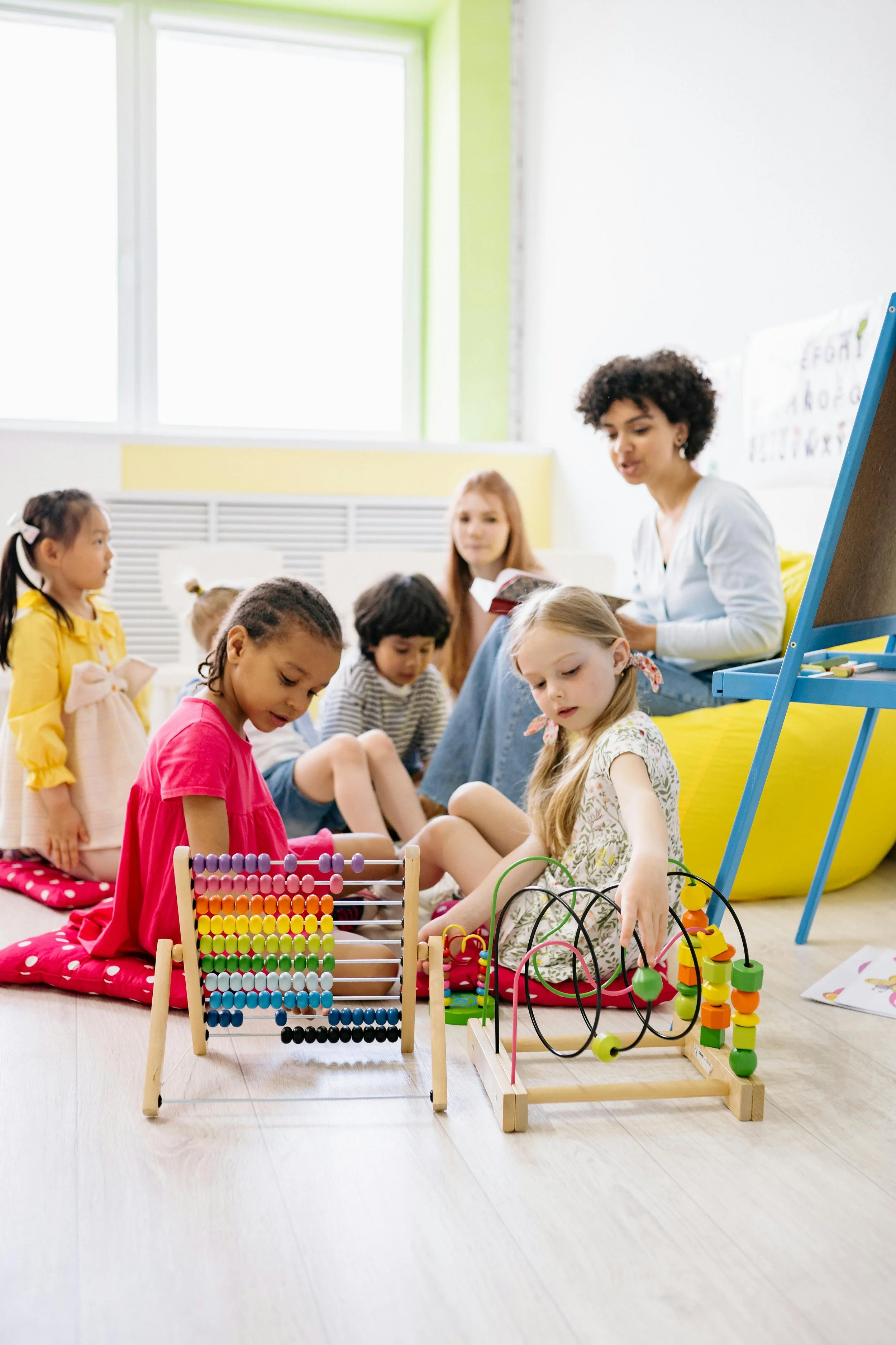 Young children sitting on the floor in a classroom, engaging with educational toys, with a teacher and a whiteboard in the background.