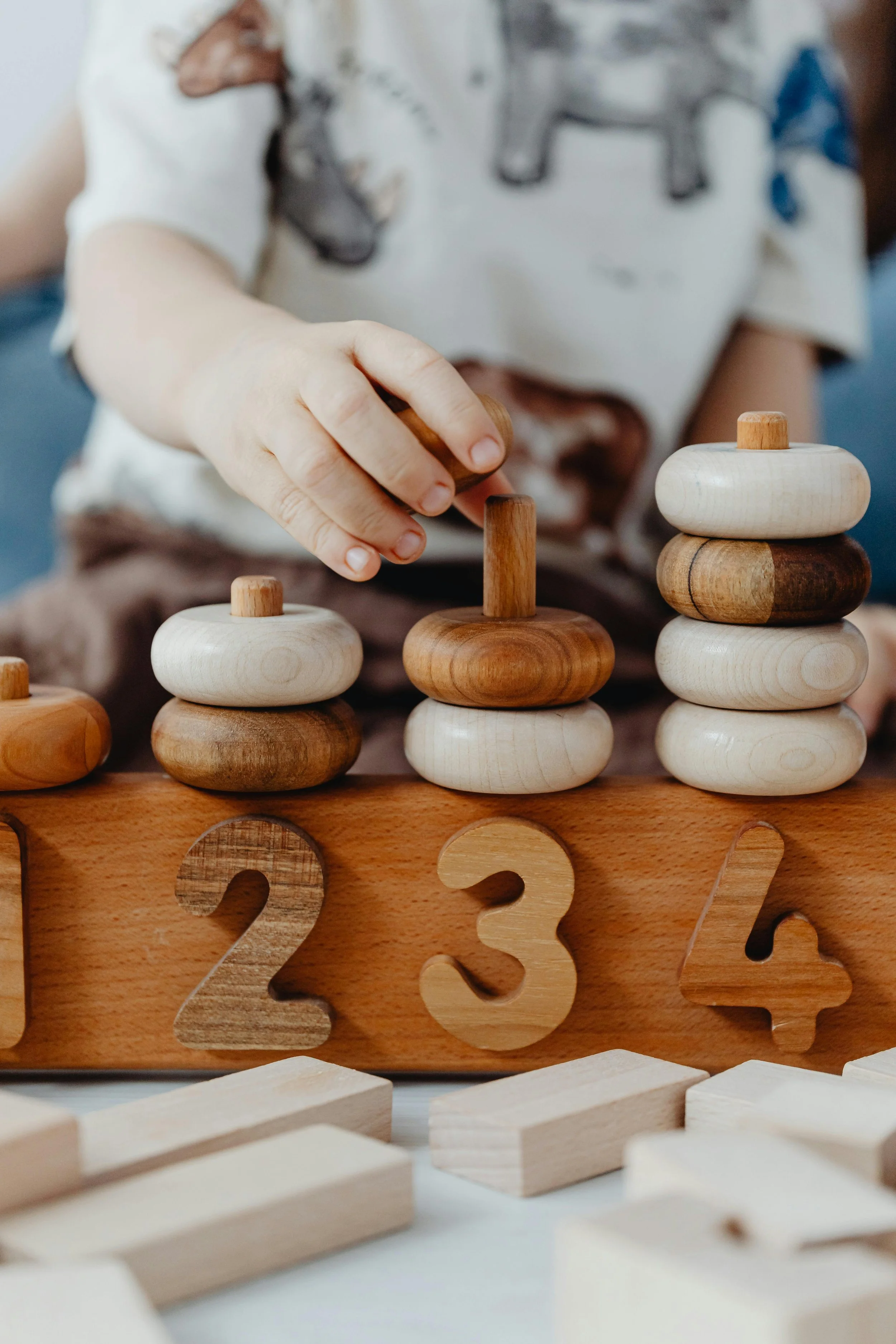 A child's hand stacking wooden animal-shaped blocks on numbered wooden base, with additional blocks and numbers in the foreground.