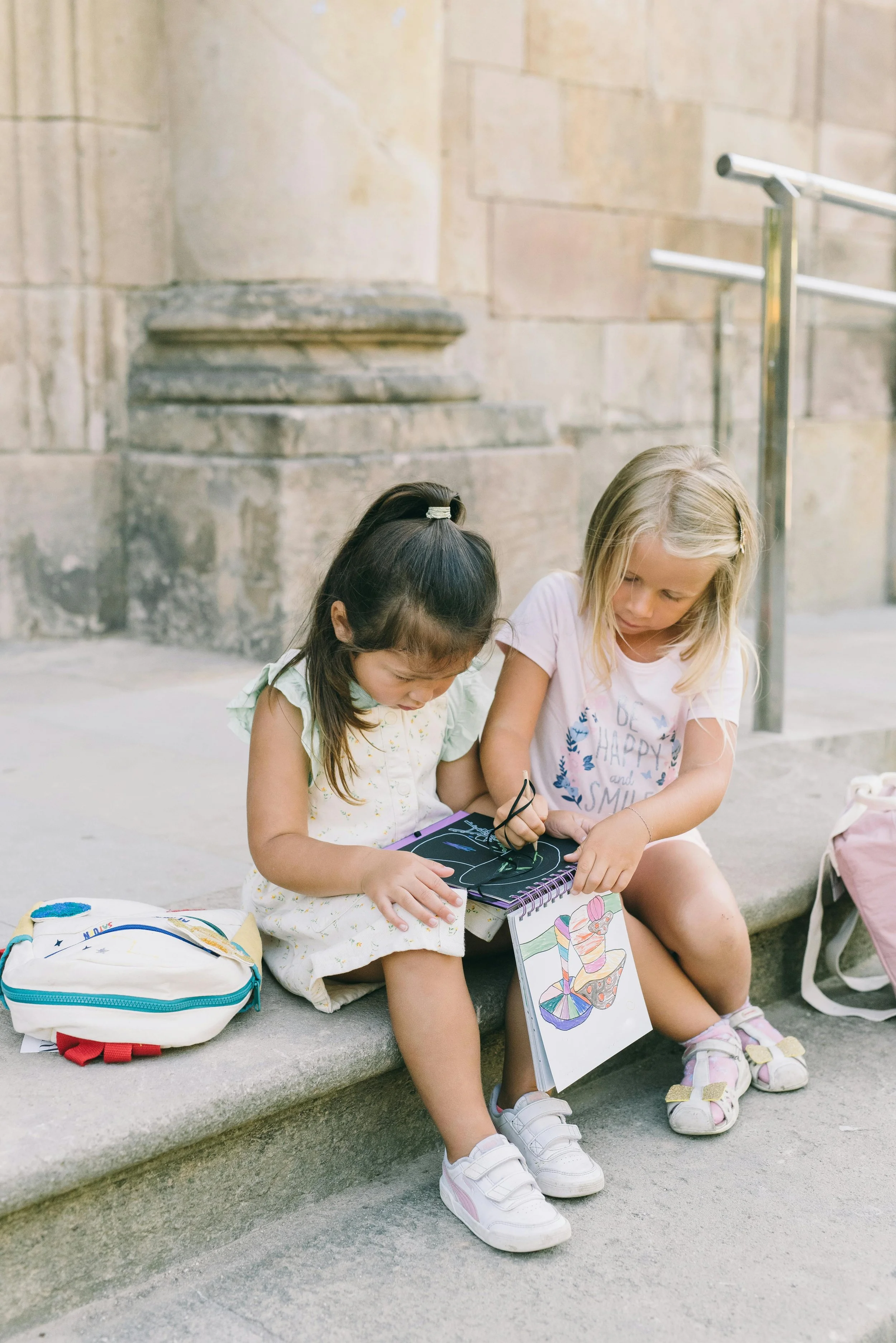 Two young girls sitting on a concrete step outside, looking at drawings and a chalkboard, with backpacks nearby.