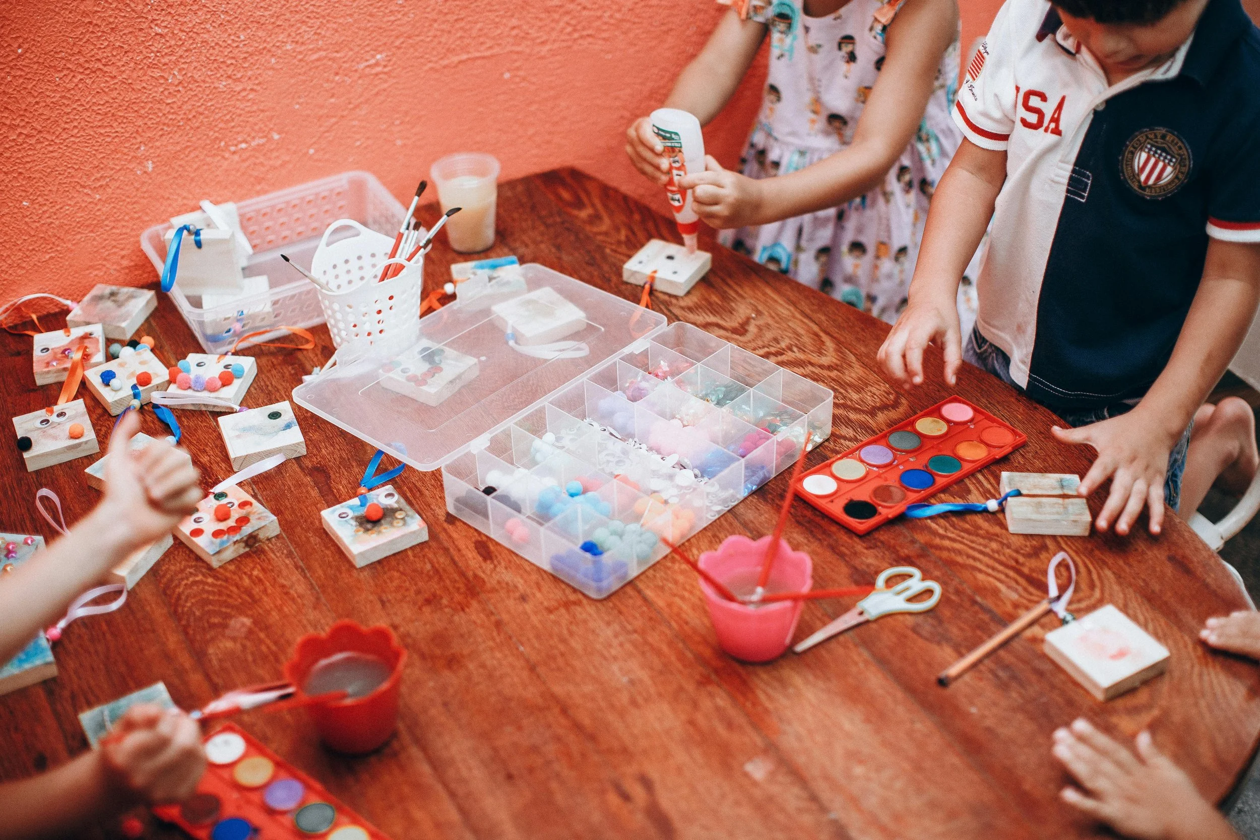 Children decorating small wooden boxes with colorful pom-poms and paint at a craft table.