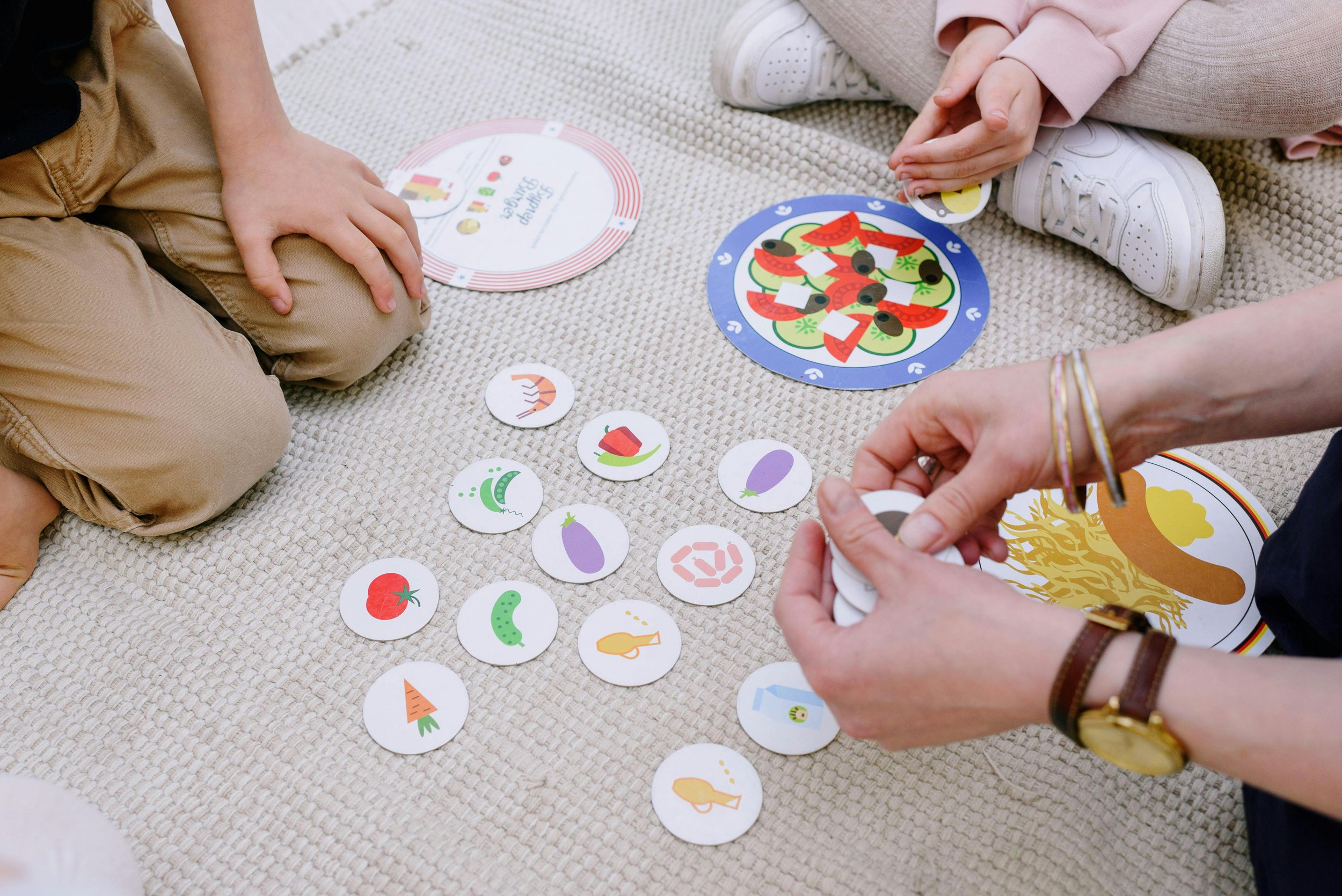 Children and an adult playing a food-themed matching game on a beige carpet, with colorful circular game pieces featuring vegetables, fish, and other food items.