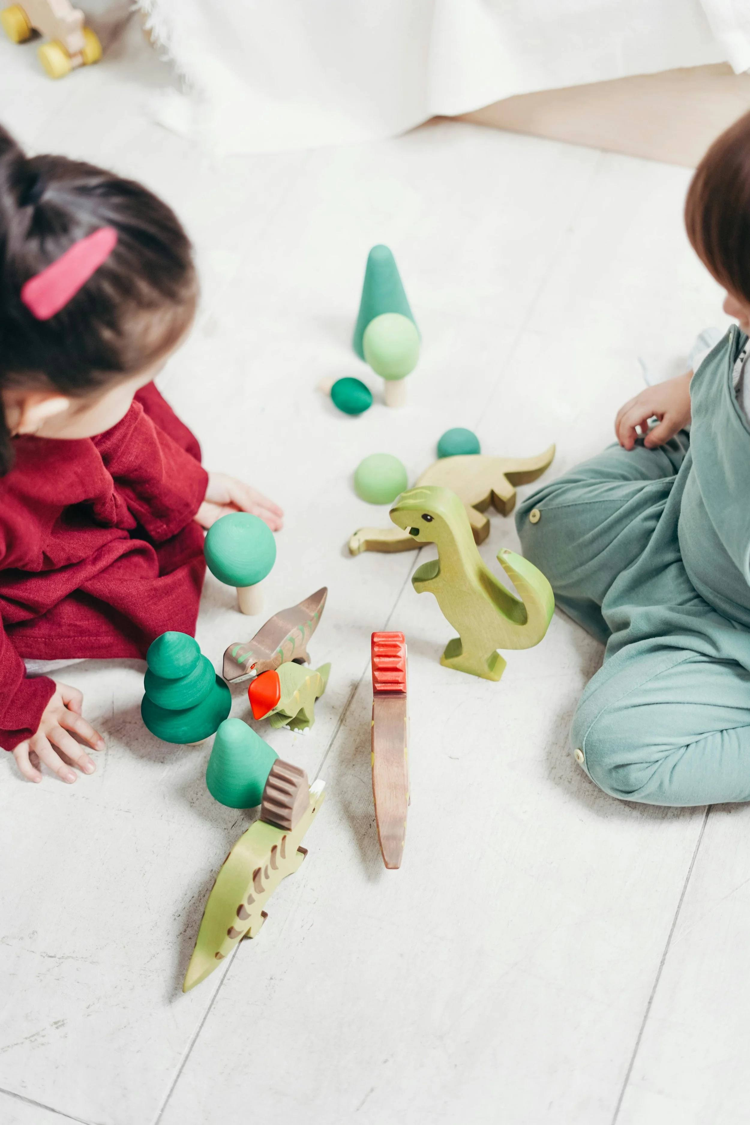 Two children playing with wooden dinosaur and tree toys on the floor.