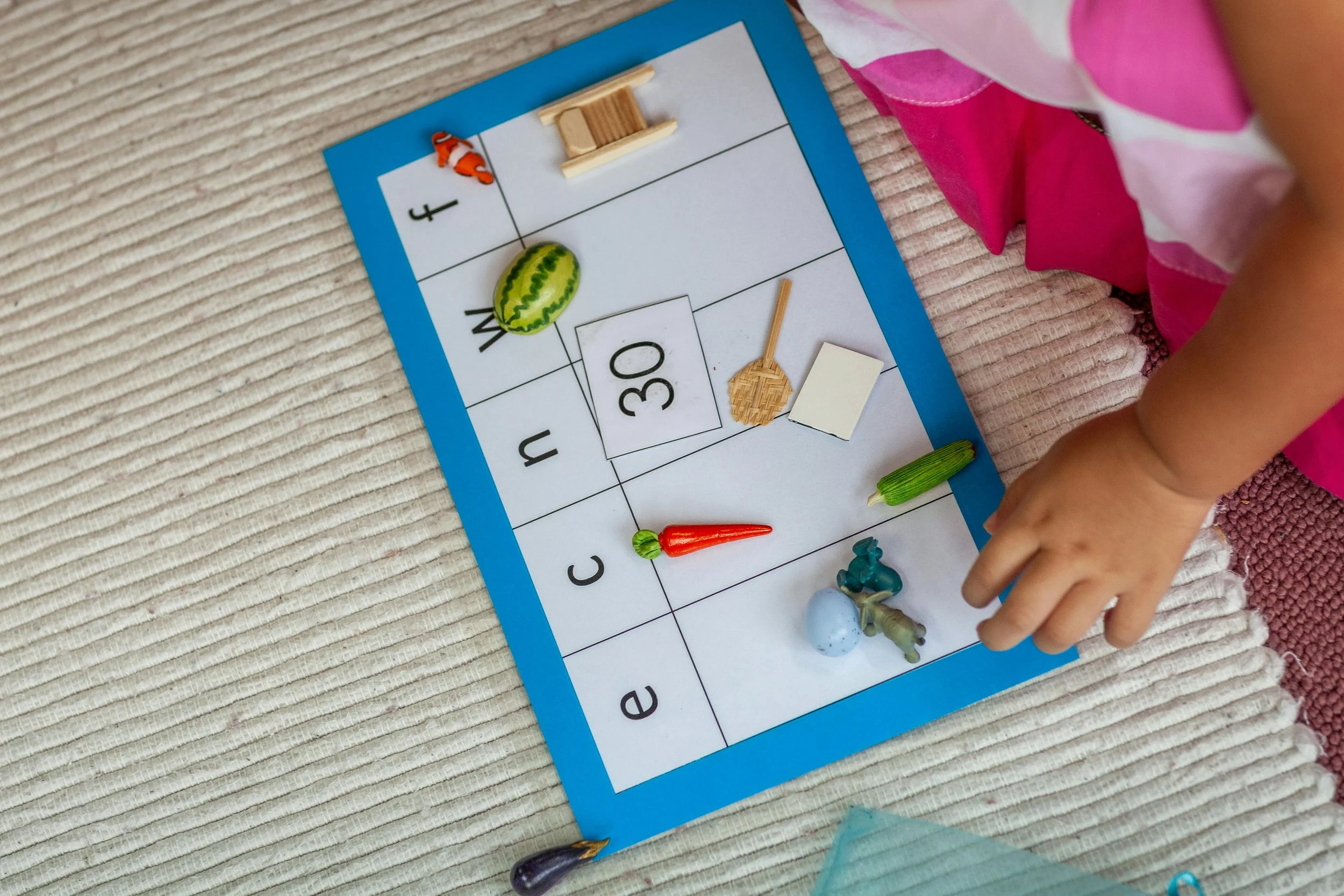 A child's hand resting near a board game with colorful plastic fish, cucumber, carrot, blue egg, and miniature objects on a woven fabric surface.