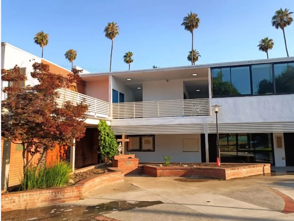 Modern building with white facade, large glass windows, balcony, and surrounding palm trees and garden areas.
