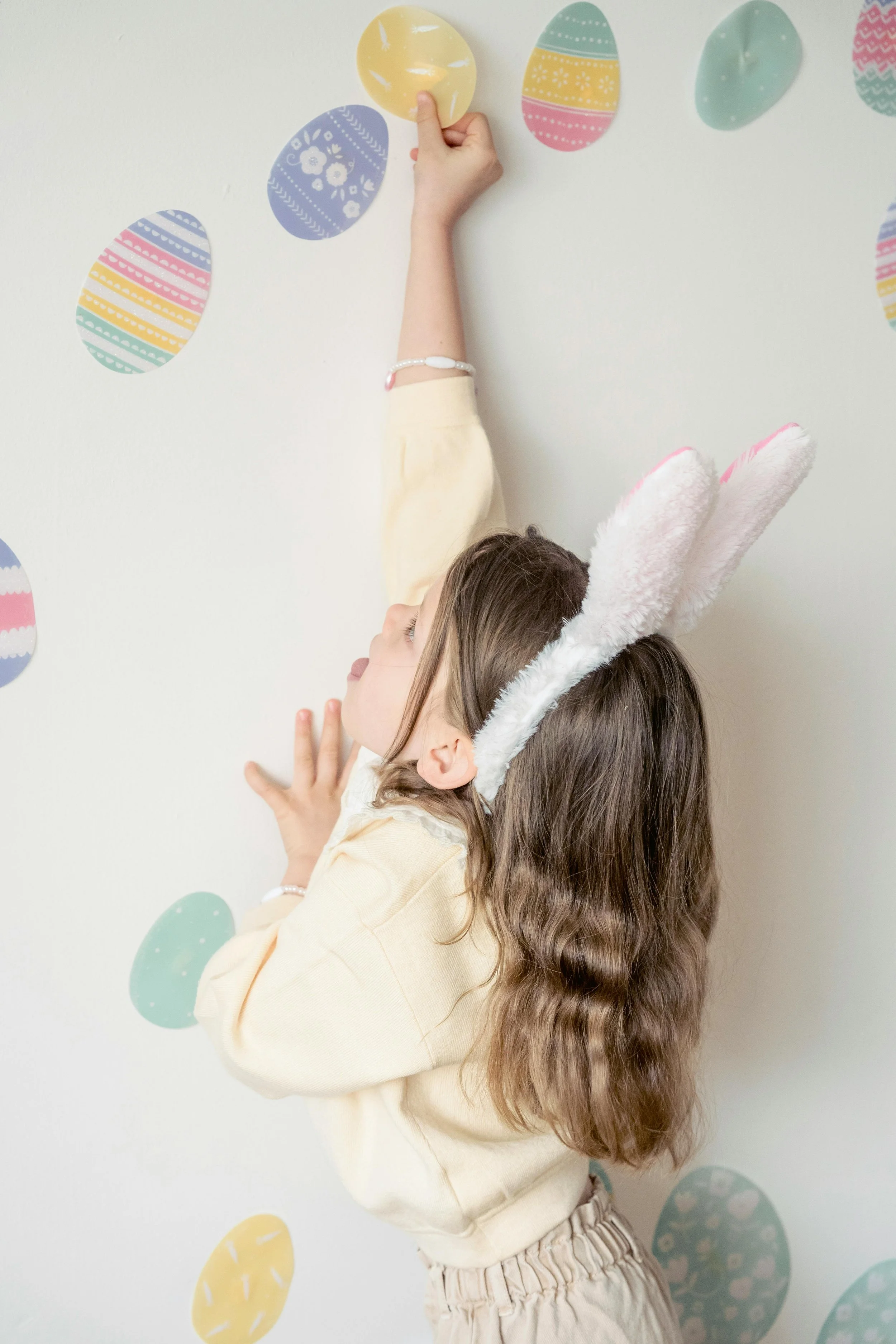 Child wearing bunny ears reaching to hang a colorful Easter egg on a wall decorated with Easter eggs