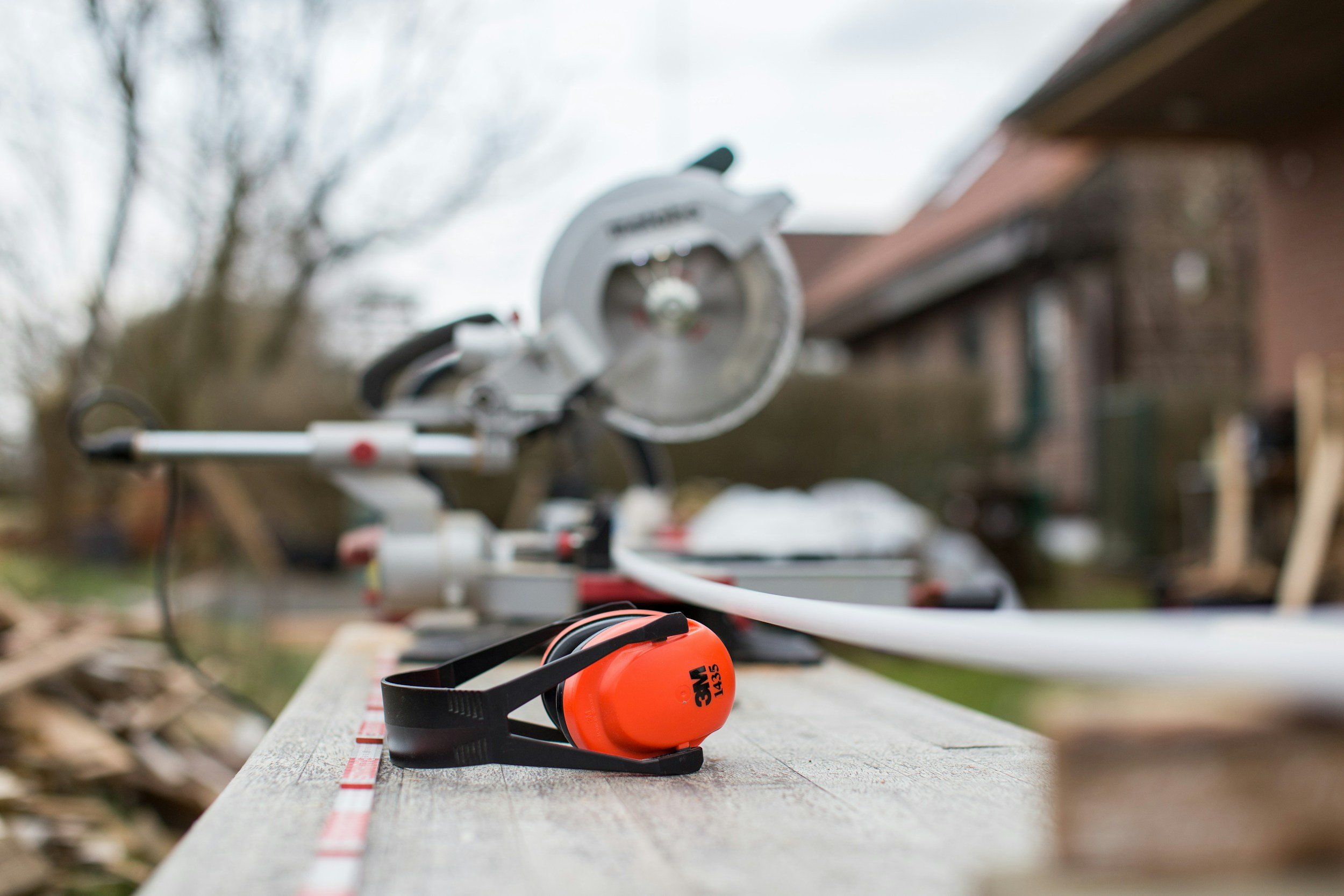 A tape measure, ear protection, and a miter saw placed on a wooden surface outdoors, with a house and trees in the blurred background.