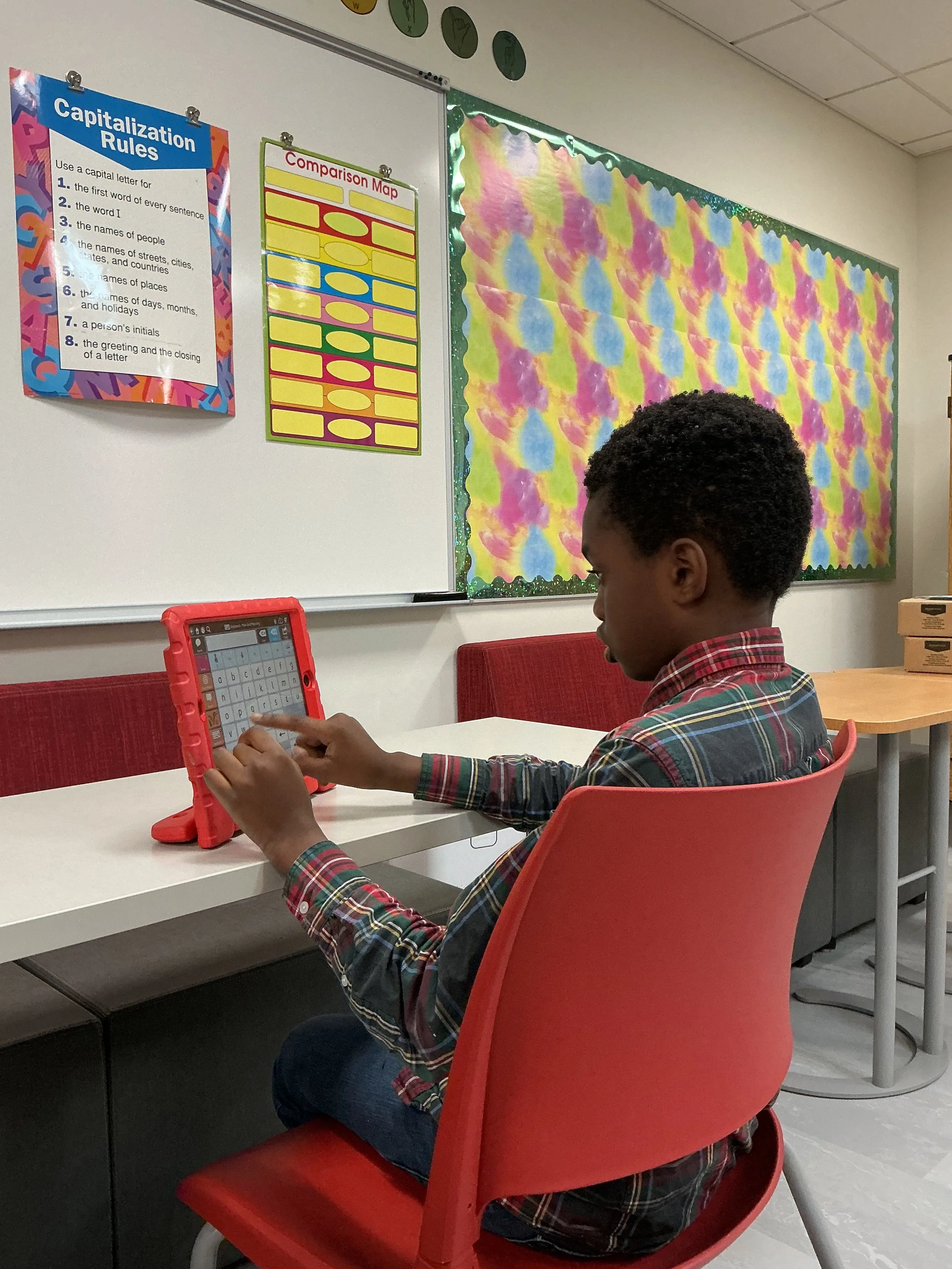 Young boy in a plaid shirt sitting in a red chair at a white desk, using a tablet in a classroom. The classroom has colorful bulletin boards on the wall, including one with capsulation rules and comparison map, and another with a abstract, tie-dye style background.