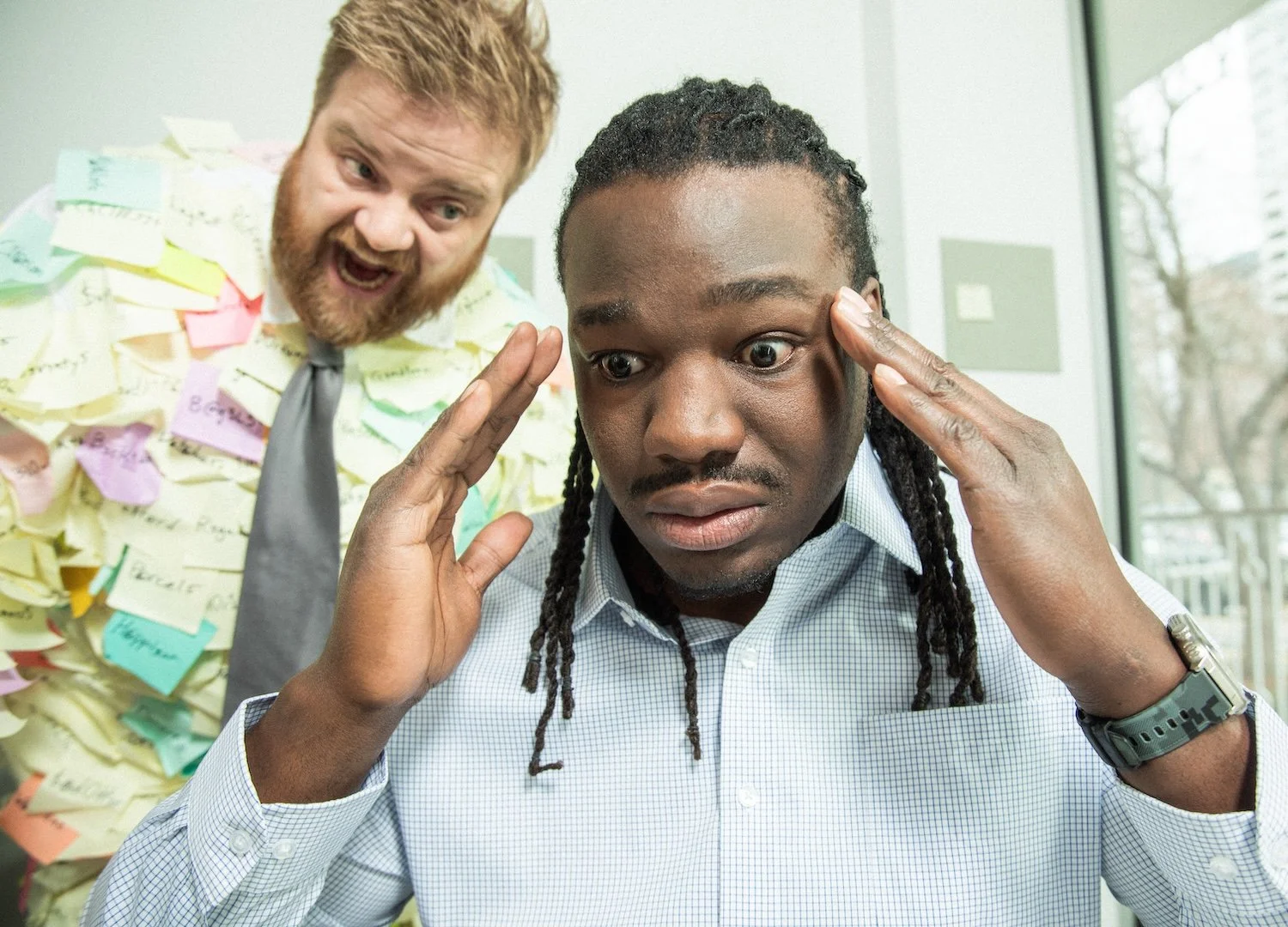 A man with long dreadlocks and a light blue shirt looks surprised as he touches his temples. Behind him, a man with red hair and a beard, wearing a shirt with numerous sticky notes, appears to be shouting or explaining something enthusiastically.
