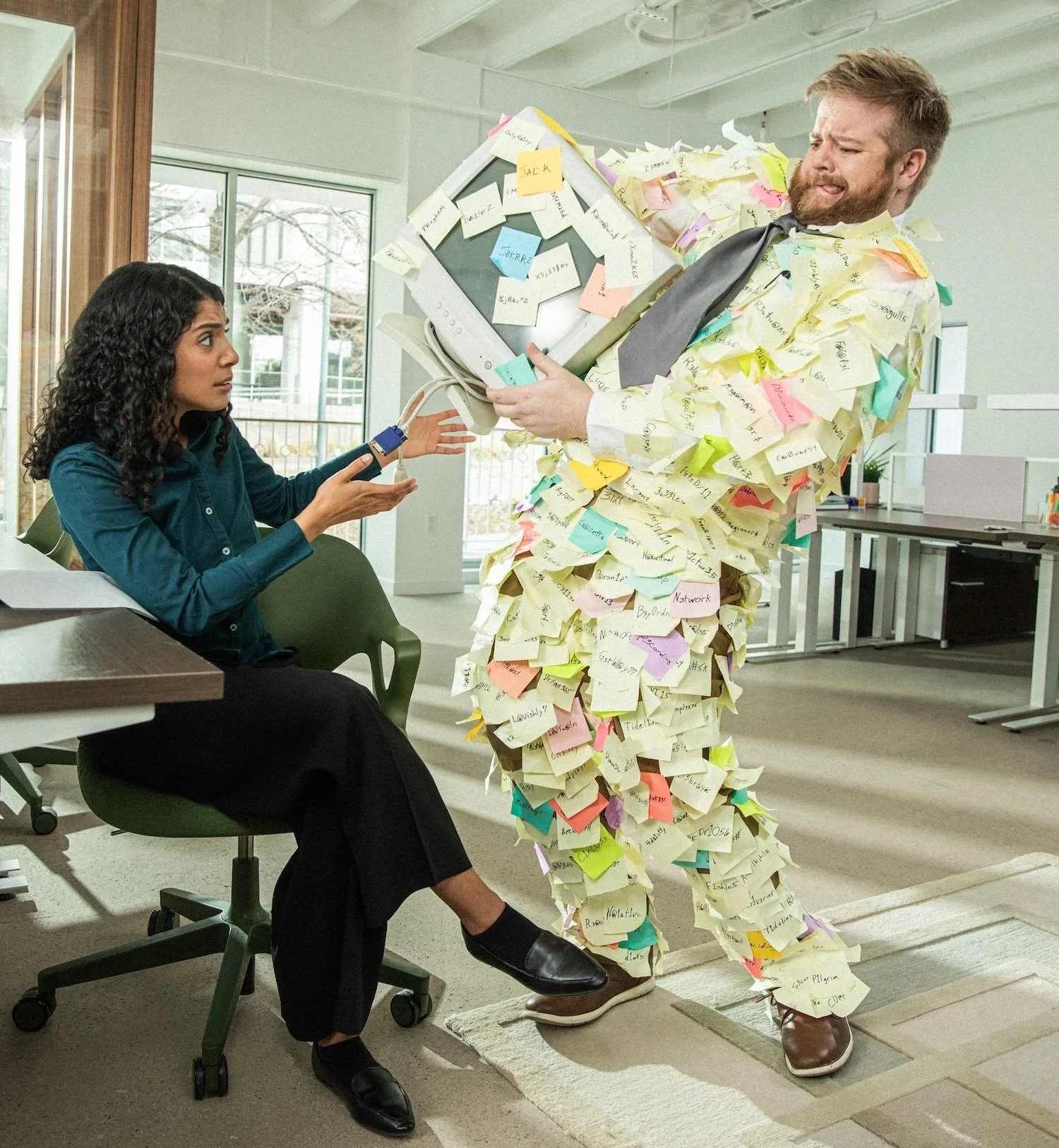 Office scene with a woman sitting at a desk and a man wearing a suit covered in sticky notes. The man is holding a computer monitor with sticky notes on it, and the woman appears to be reaching out to him with a frustrated expression.