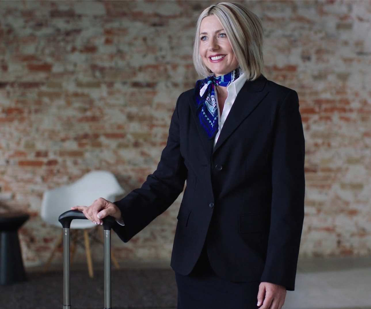 A smiling woman with blonde hair in a black business suit, standing in front of a brick wall, holding a suitcase handle, wearing a blue patterned scarf.