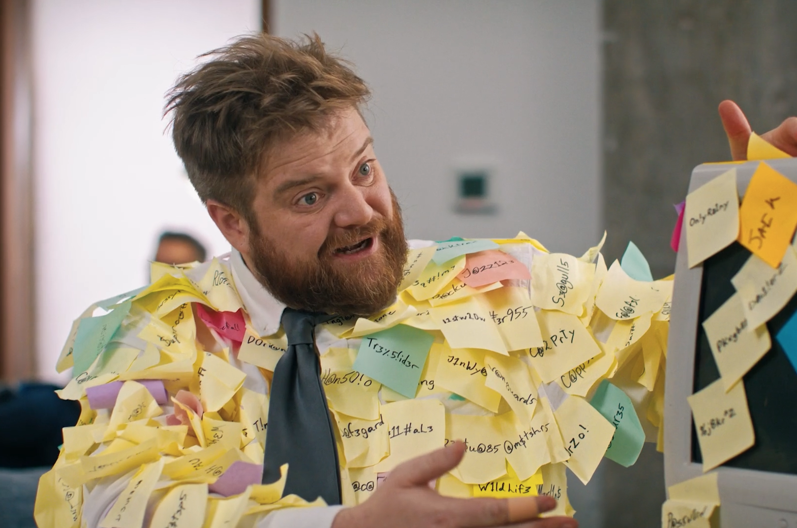 A man with red hair and beard, wearing a white shirt, grey tie, and covered with yellow, pink, and green sticky notes, standing in front of a computer monitor in an office, appearing excited or surprised.