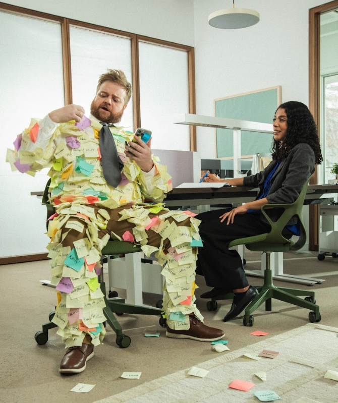A man in an office, covered in colorful sticky notes, sitting on a chair while a woman at her desk looks on, smiling.