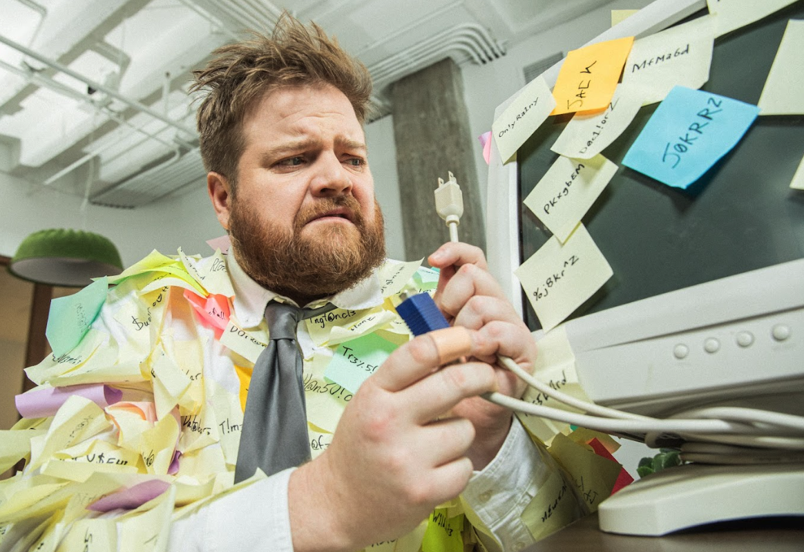 A man with a beard and messy hair, wearing a shirt, tie, and covered with numerous colorful sticky notes, looks frustrated while working on a computer in an office environment.