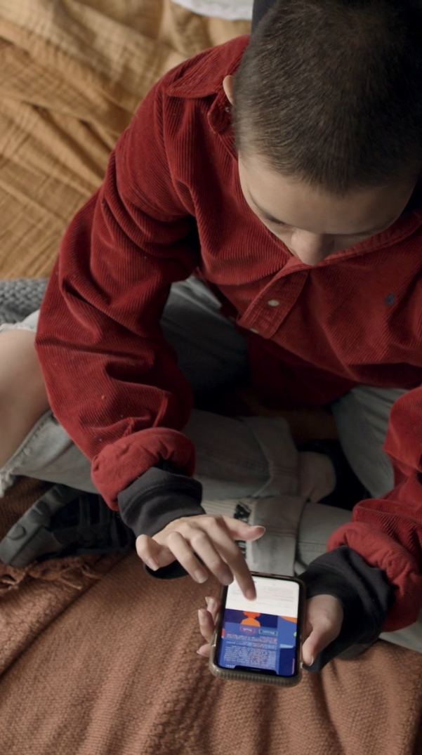 A boy sitting on a couch, wearing a red jacket and holding a smartphone, looking at the screen.