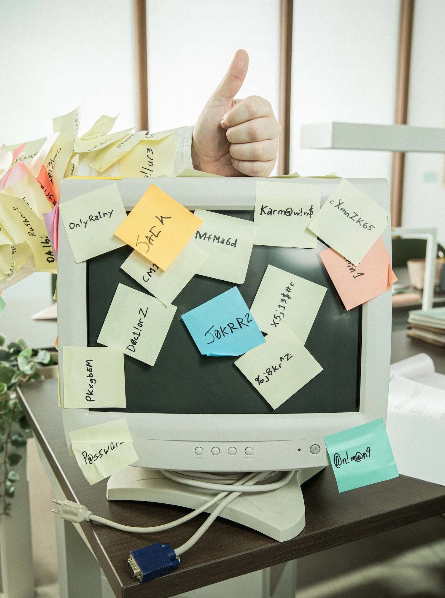 Computer monitor covered with various sticky notes with handwritten passwords and emails, with a thumb-up gesture seen behind the monitor.