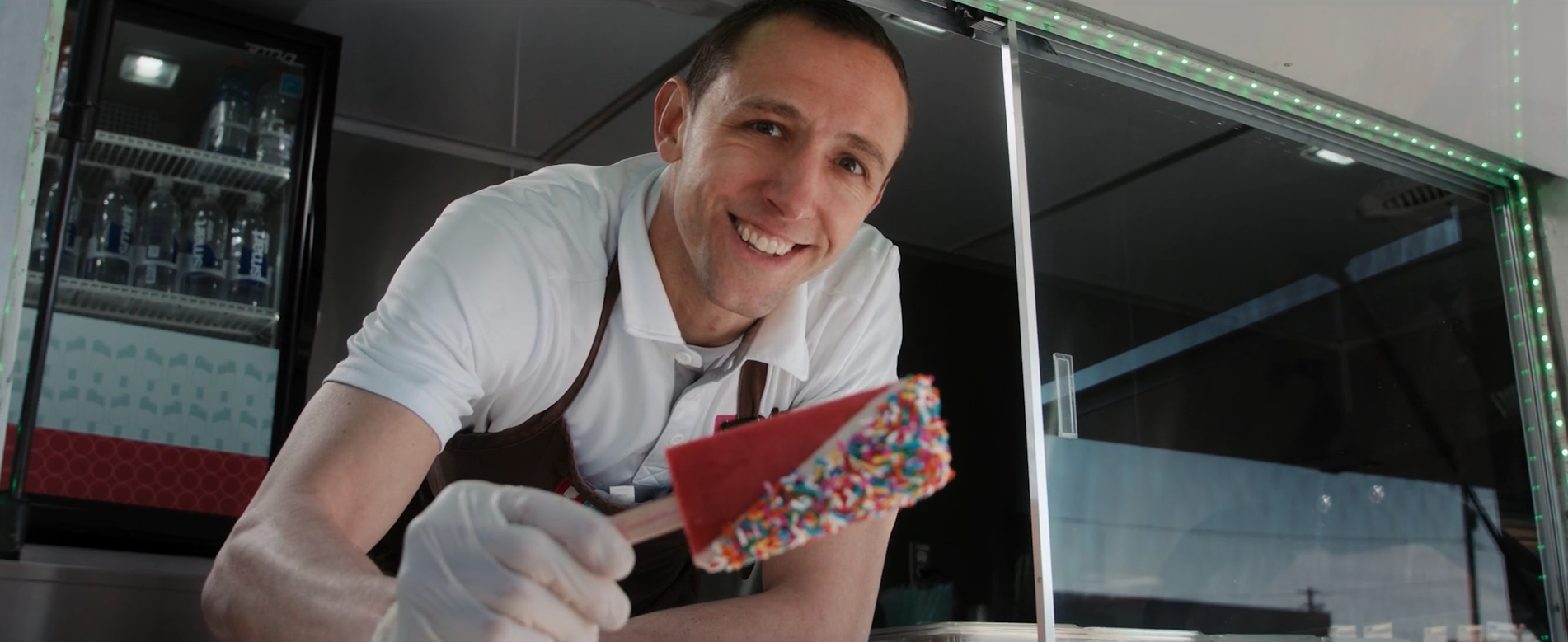 A man smiling and looking at the camera while holding an ice cream cone with colorful sprinkles, inside an ice cream truck.