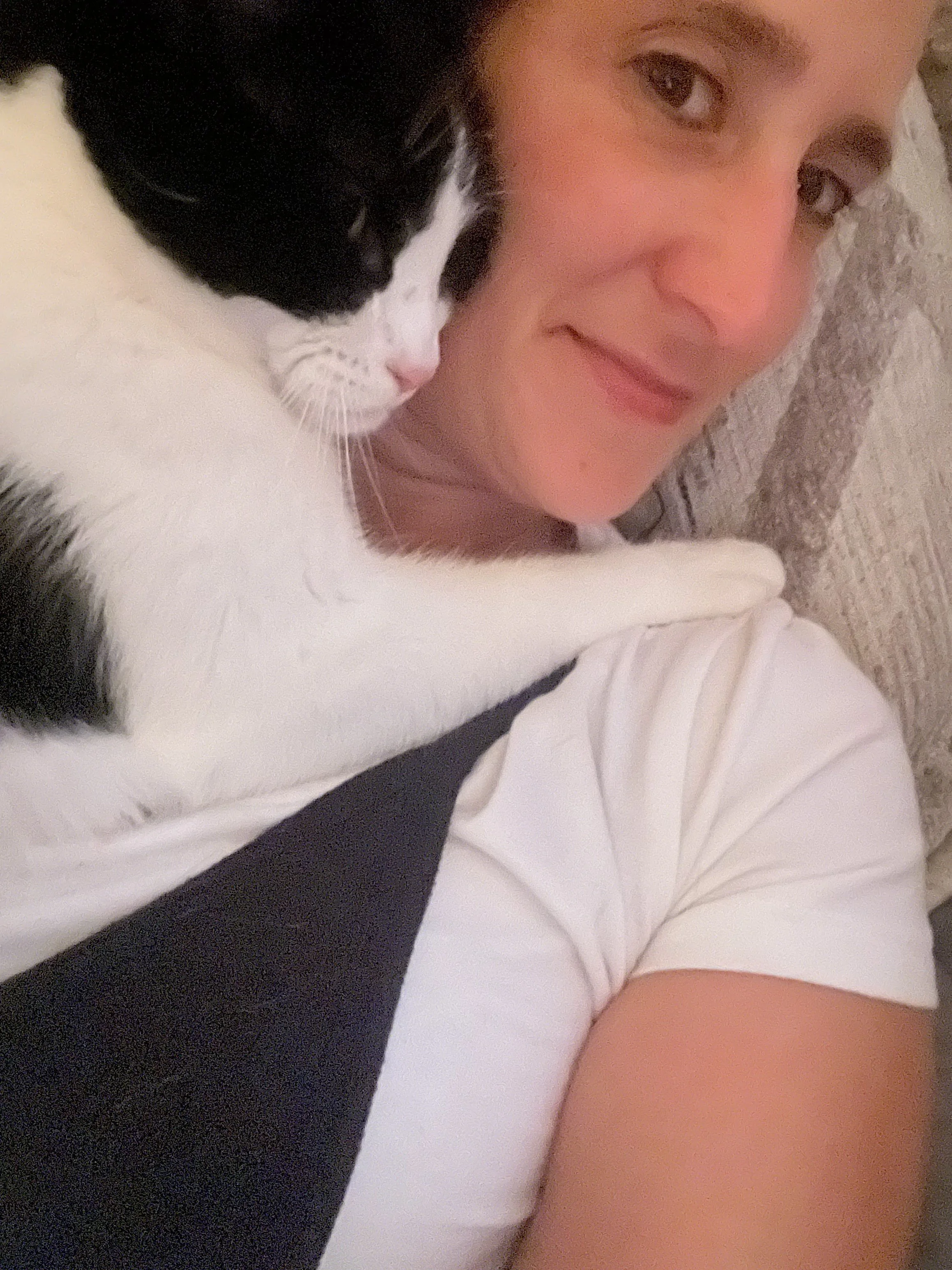 A woman lying on a couch with a black and white kitten resting on her shoulder and neck