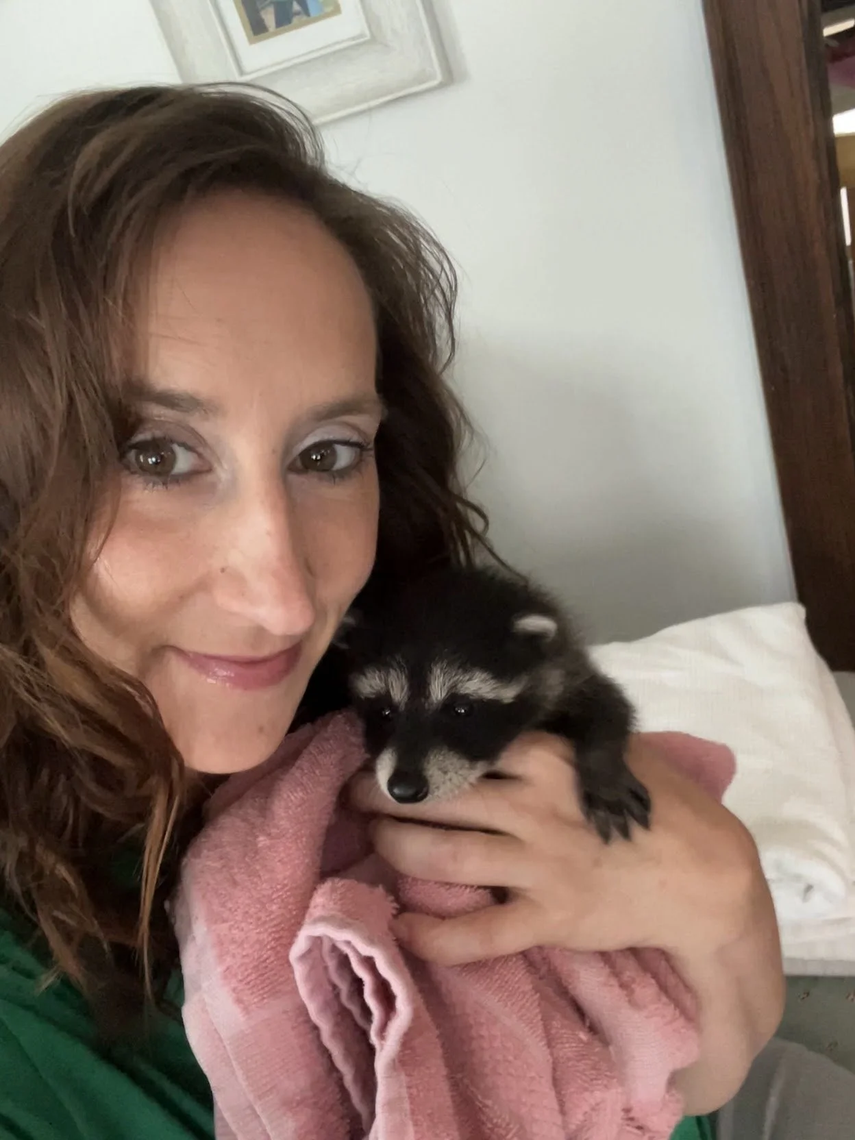 A woman with curly brown hair holding a small black and white puppy in her arms, wrapped in a pink towel, indoors with a bed and framed picture visible in the background.