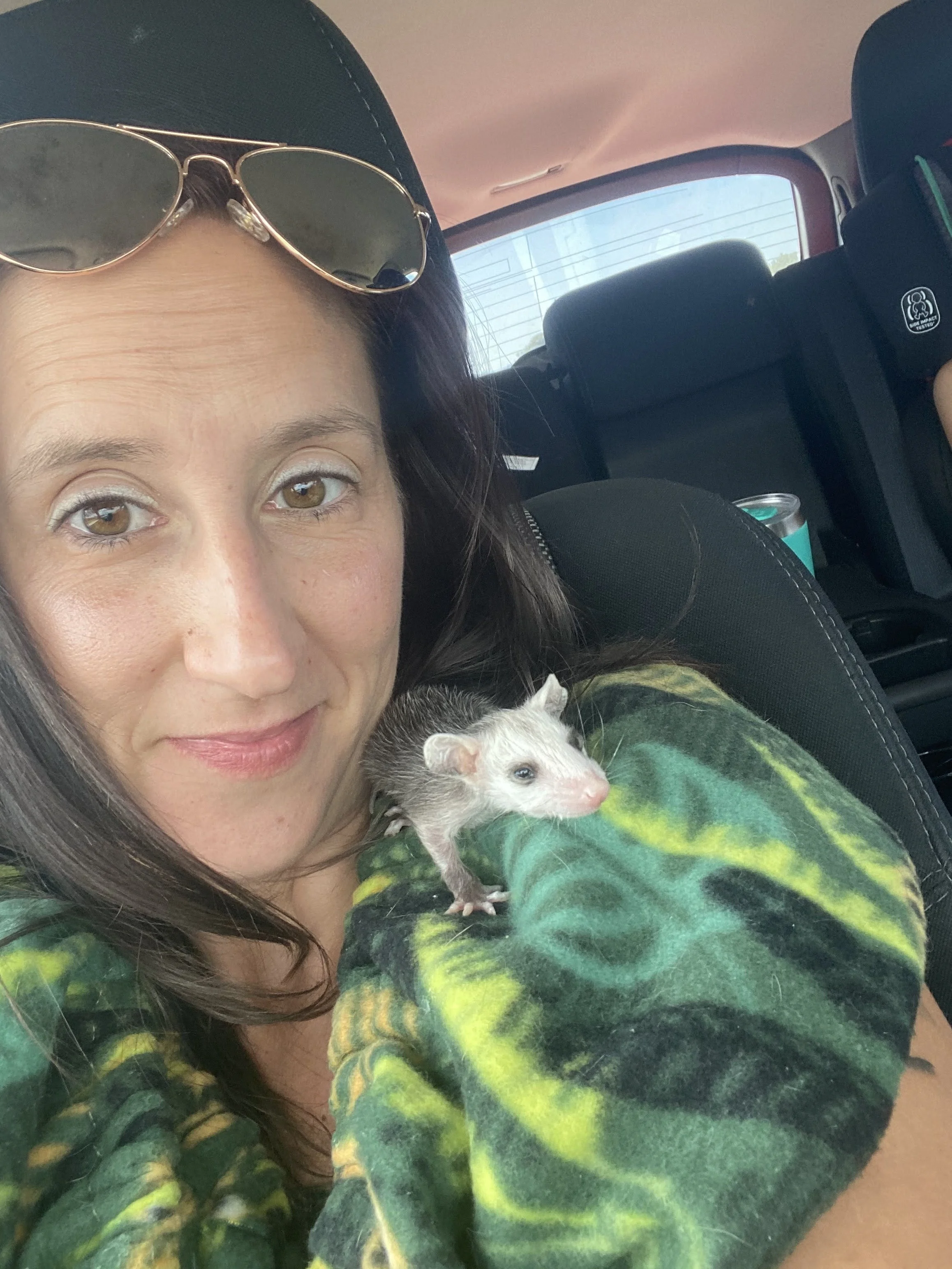 A woman with brown hair and light makeup holding a small ferret in a car. She is wearing a tie-dye shirt with green and yellow colors and sunglasses on her head. There is a soda can in the backseat.