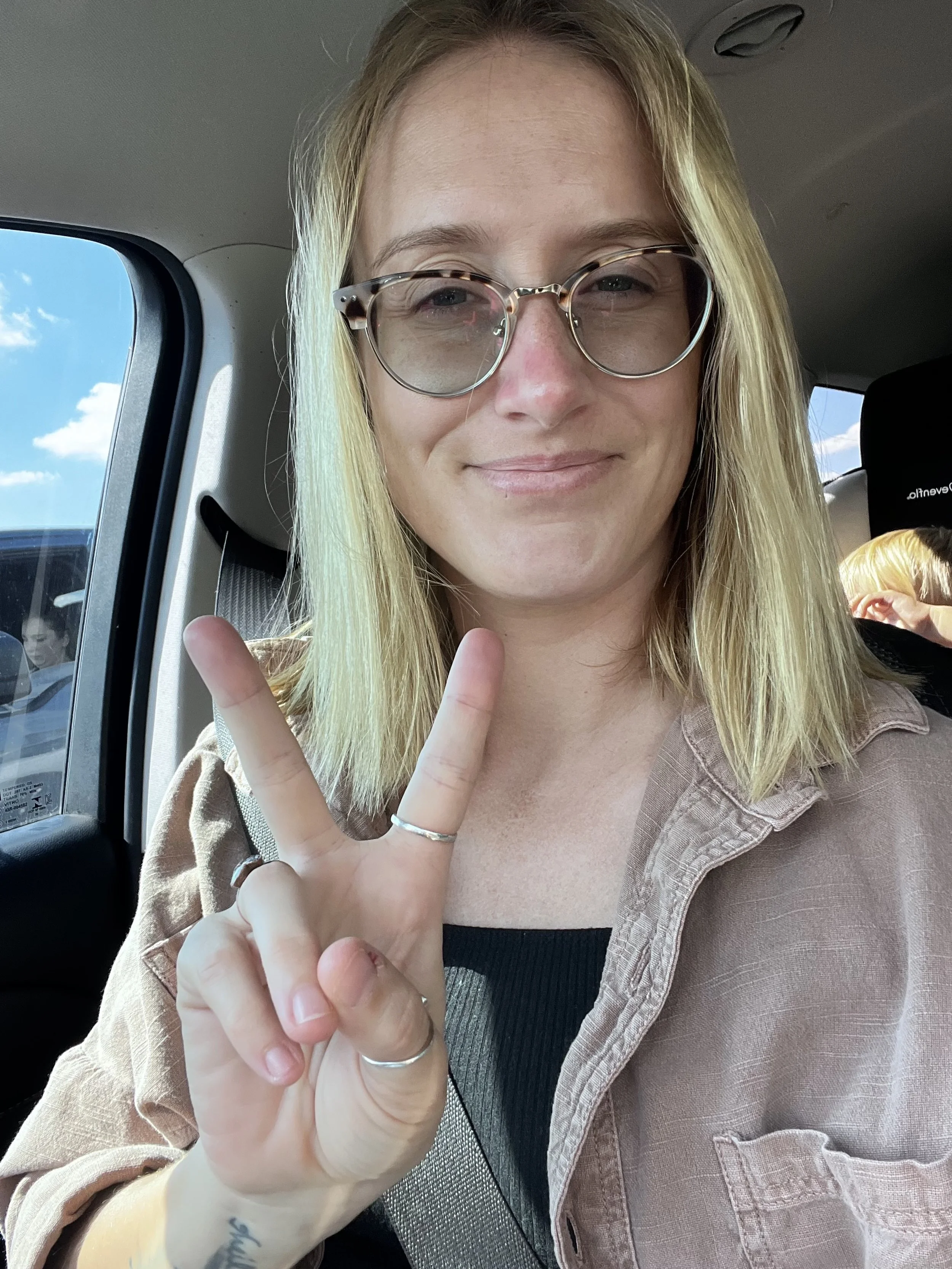 Young woman with blonde hair, glasses, and a neutral expression, making a peace sign inside a vehicle with a blue sky visible through the window.
