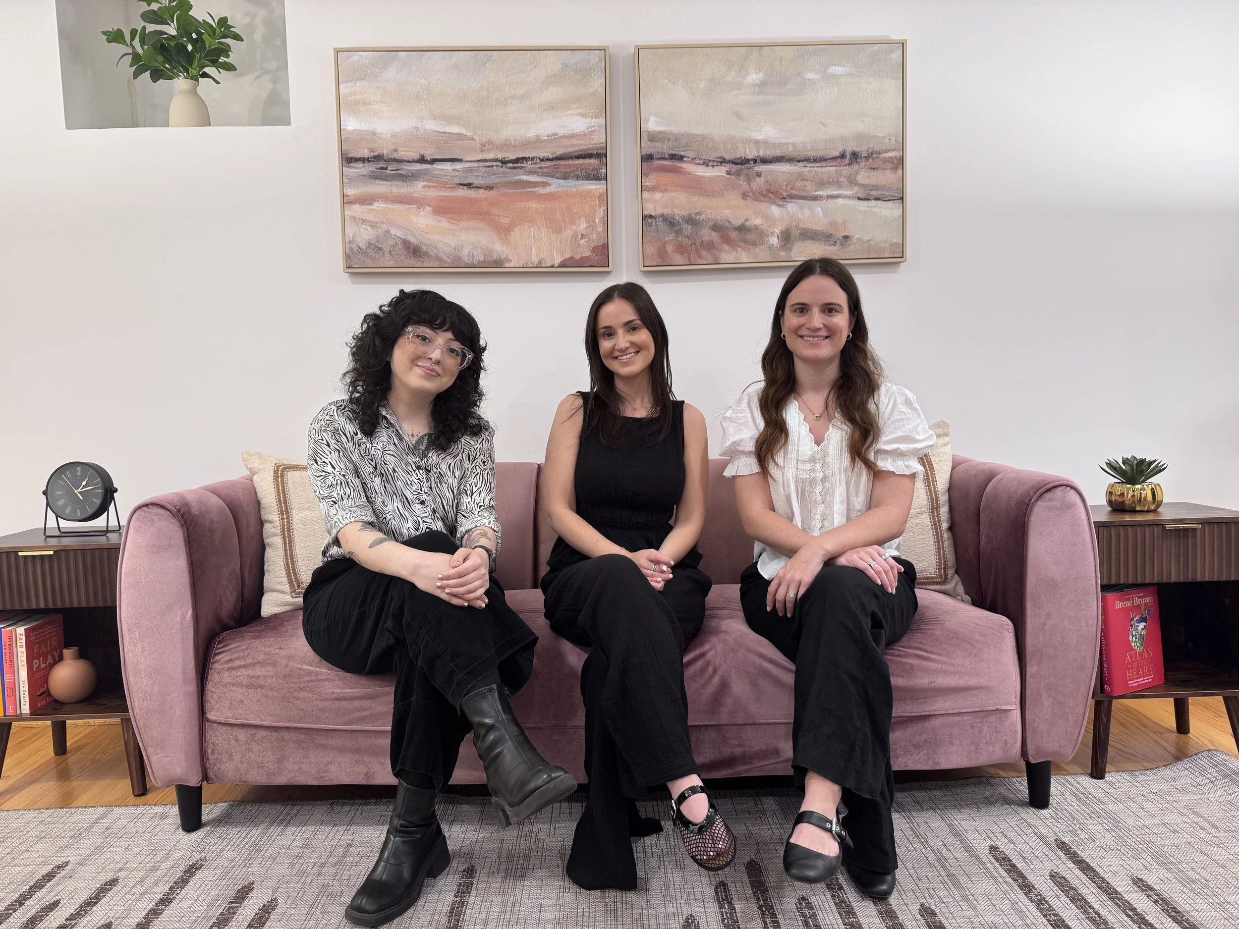 Photograph of three women sitting on a pink sofa in a living room, smiling at the camera. Behind them are two framed landscape paintings, a white wall, and a small potted plant on a side table. On the left side, there is a black alarm clock and a beige vase on a side table, and on the right side, a small plant in a gold pot and some books are visible.