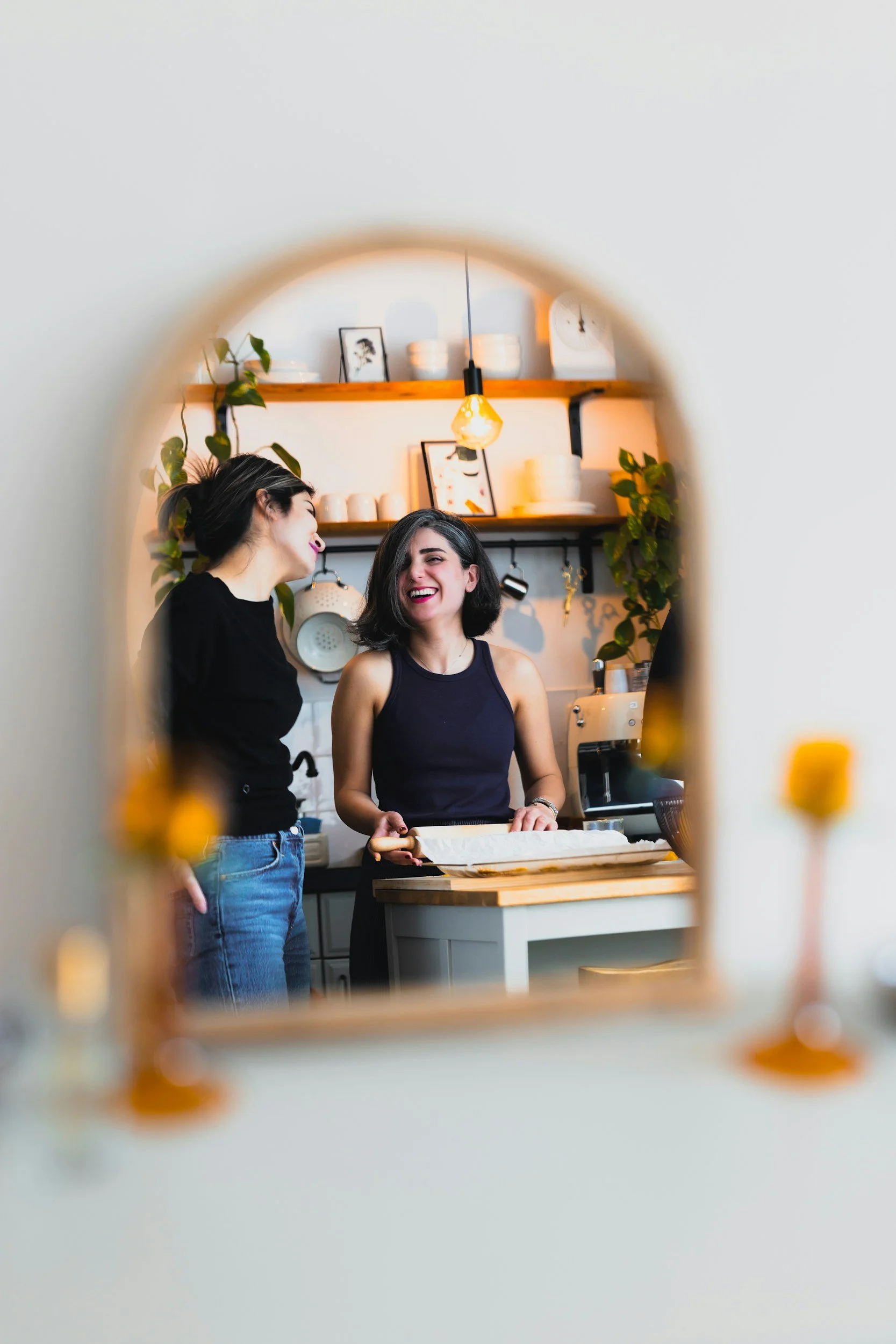 Two women smiling in a kitchen mirror reflection, representing LGBTQ-inclusive couples counseling focused on connection and honest communication.
