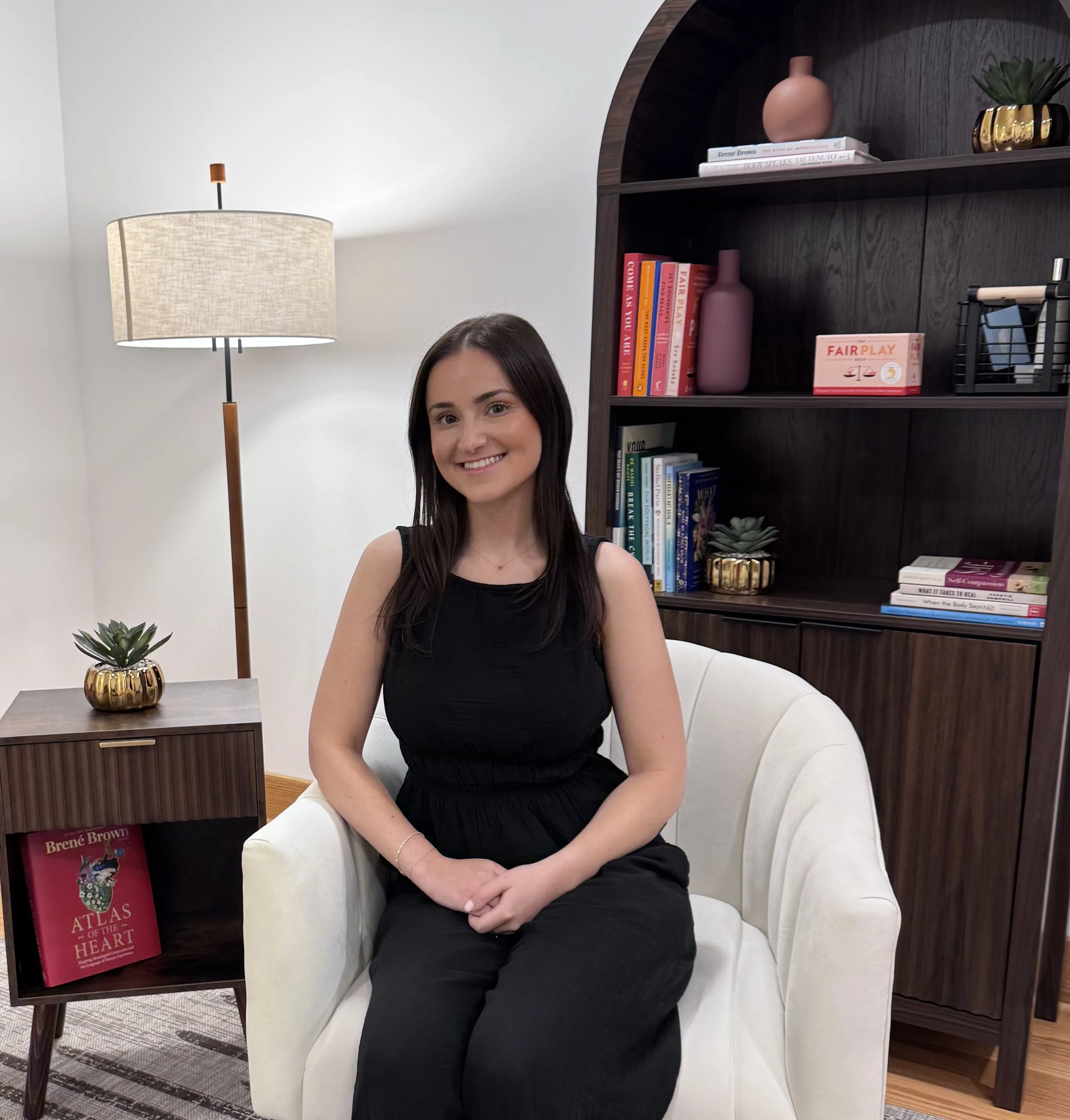 A woman with long dark hair smiling and sitting on a white chair in a room with a bookshelf, lamp, and plants.