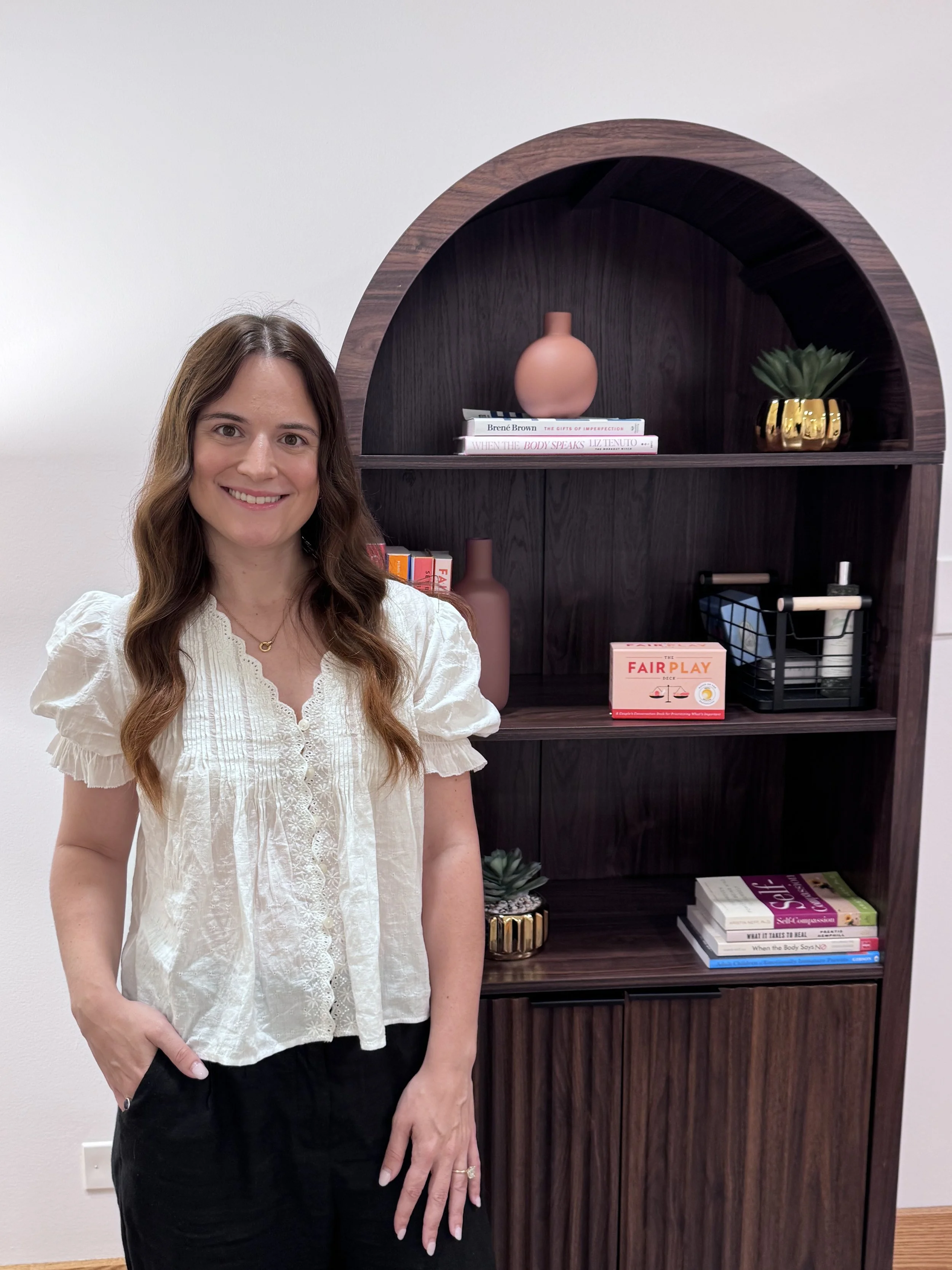 A woman with long brown hair, wearing a white blouse, standing next to a dark wood bookshelf with decorative items and books.