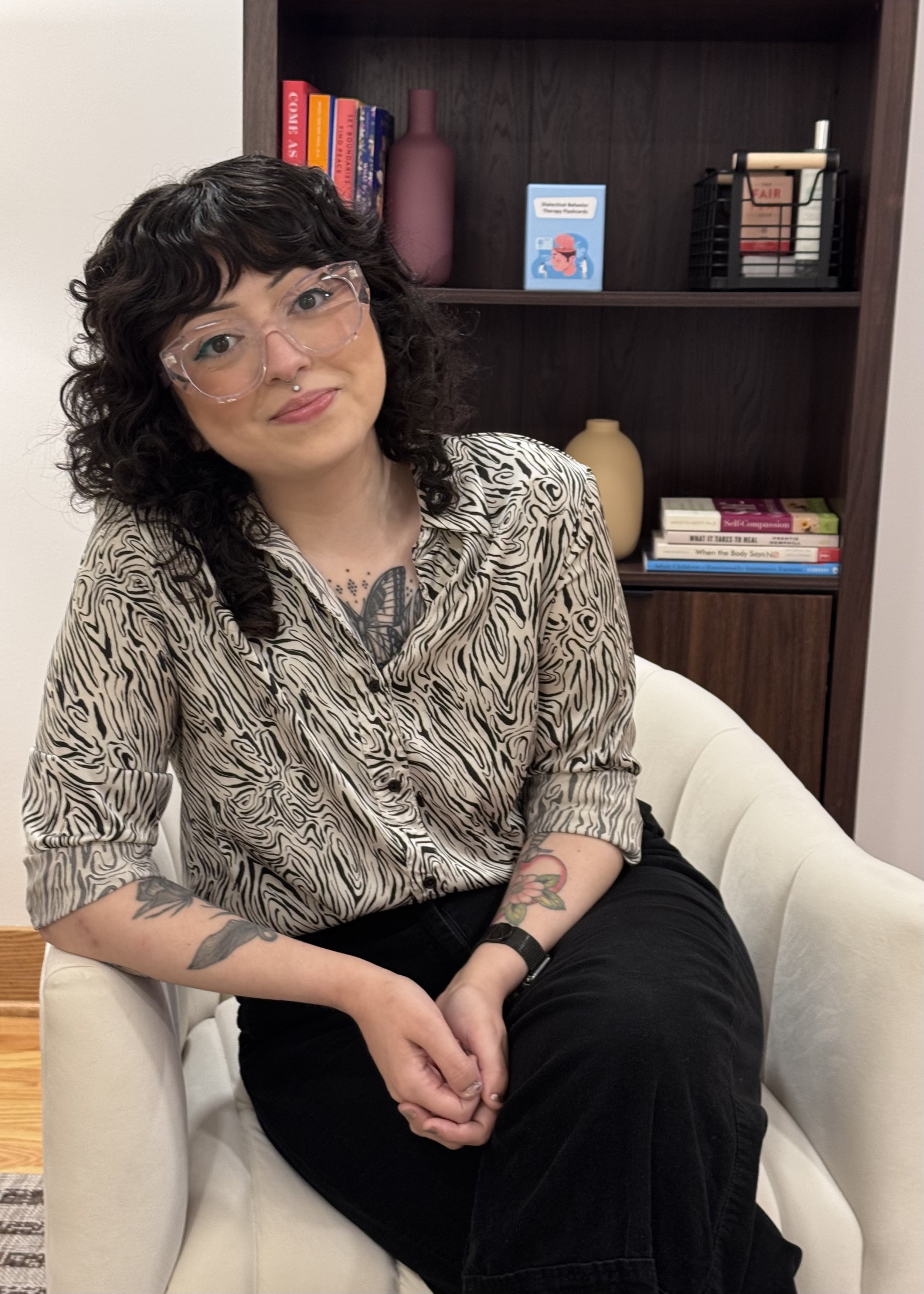 A woman with curly dark hair wearing large clear glasses, a nose piercing, and a zebra print shirt, sitting on a white armchair with her hands clasped, in a room with a wooden bookshelf behind her filled with books and decorative items.
