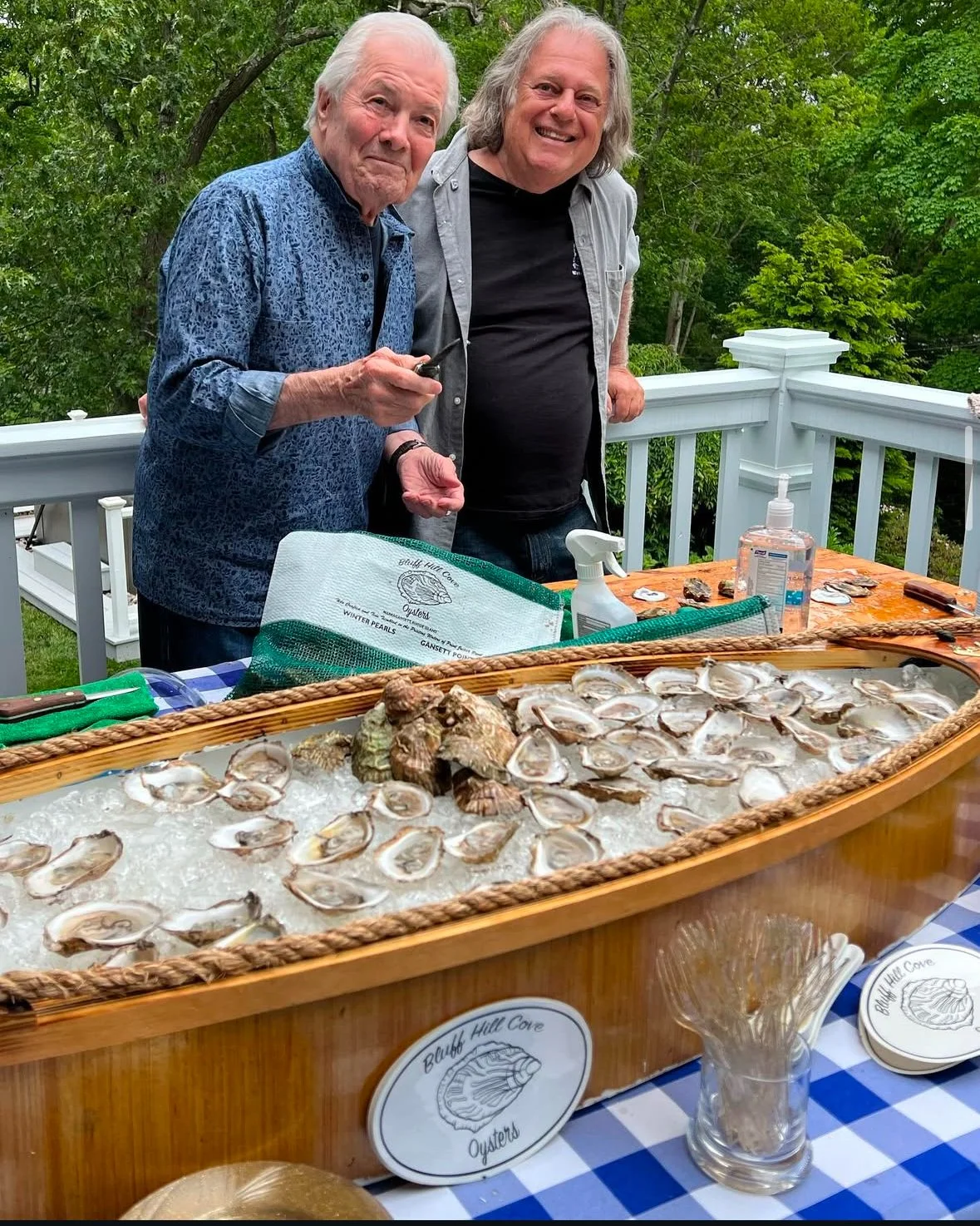 Two men standing behind a table with harvested oysters on ice, at an outdoor event, with trees in the background.