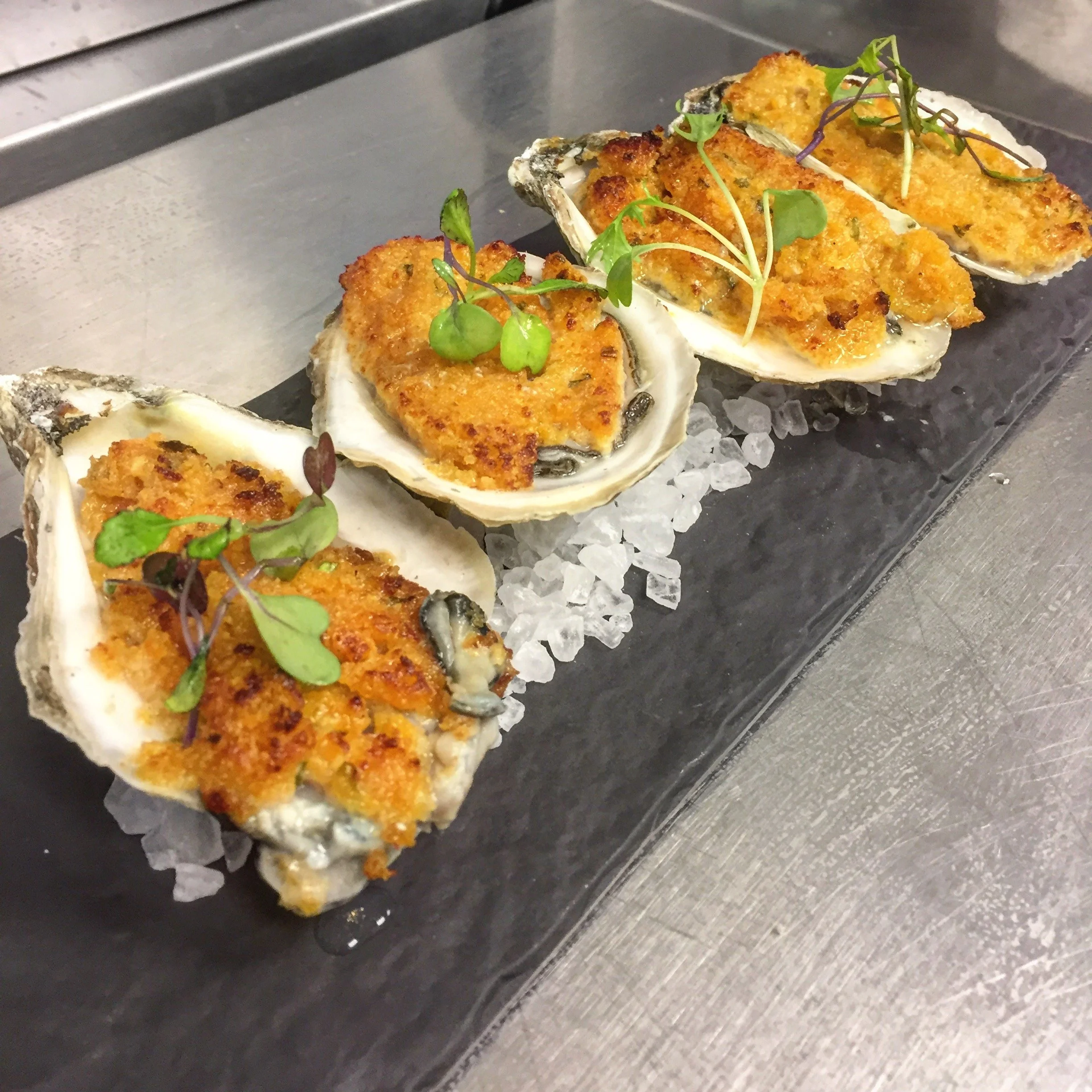 Four baked oysters topped with golden breadcrumbs and herbs, garnished with microgreens, served on a bed of crushed ice on a black slate platter.