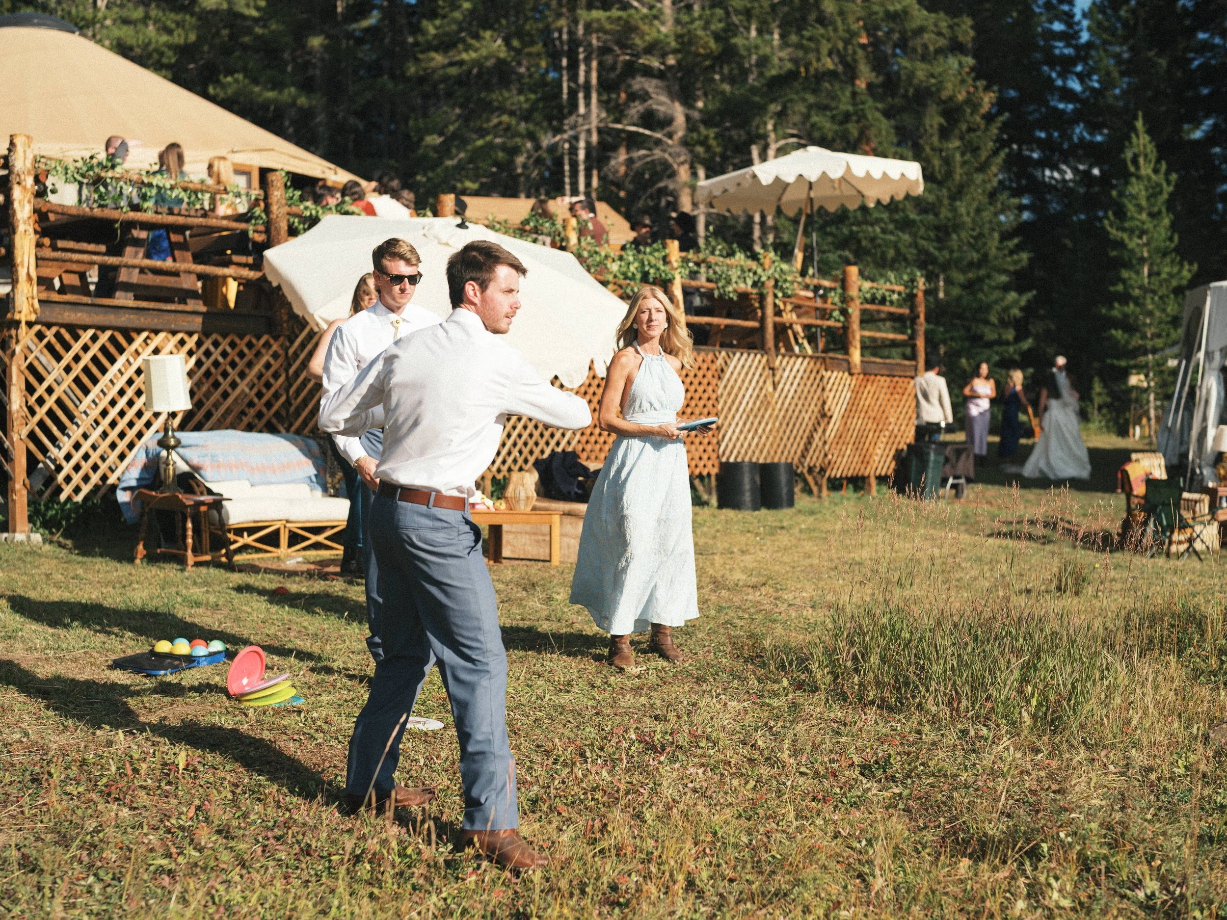 People playing a game outdoors, with colorful balls and equipment on the grass, at a gathering near tents and a wooden deck with trees in the background.