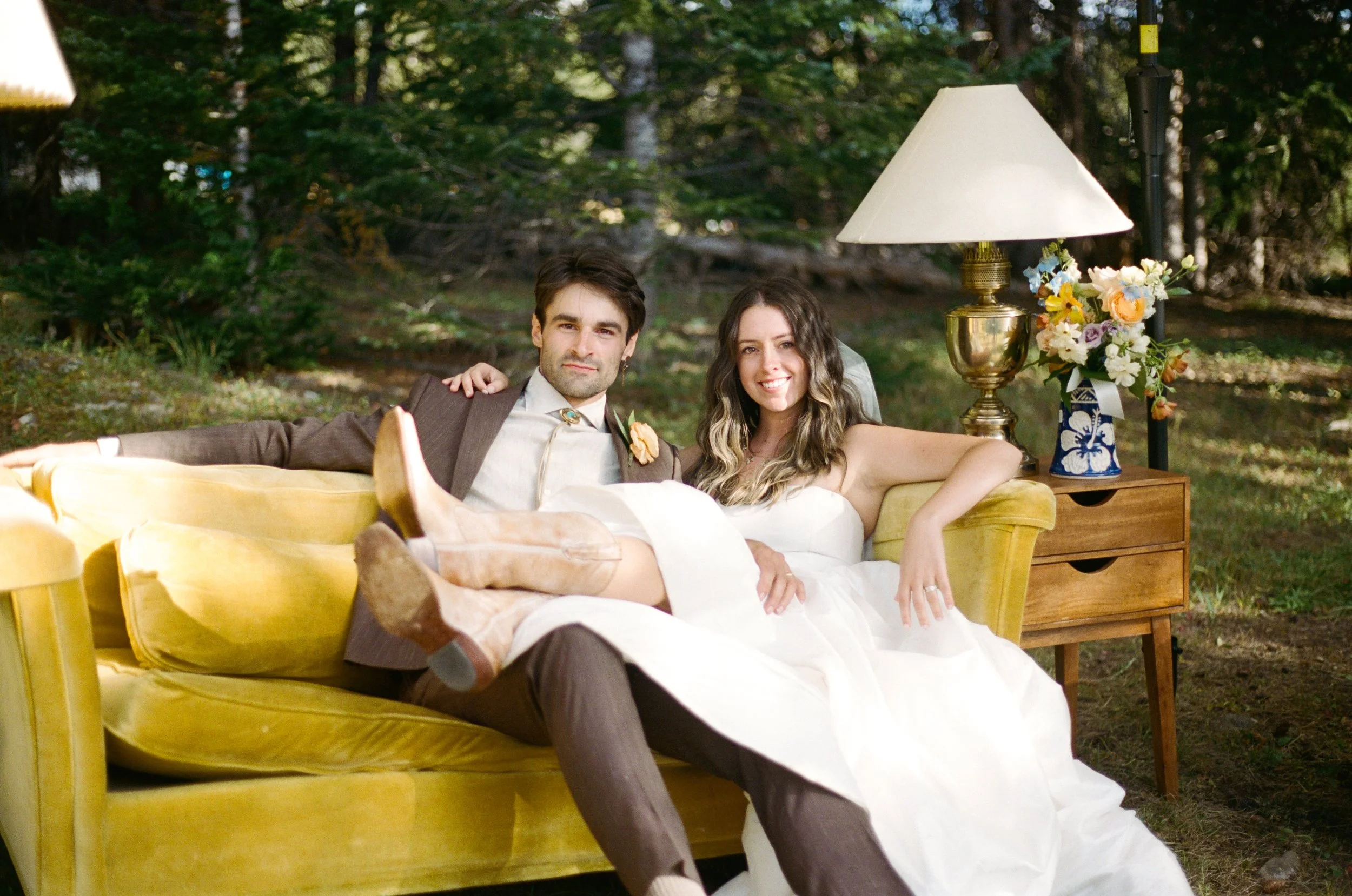 A man and woman sitting on a yellow sofa outdoors in a wooded area with a lamp and flowers beside them, smiling at the camera.