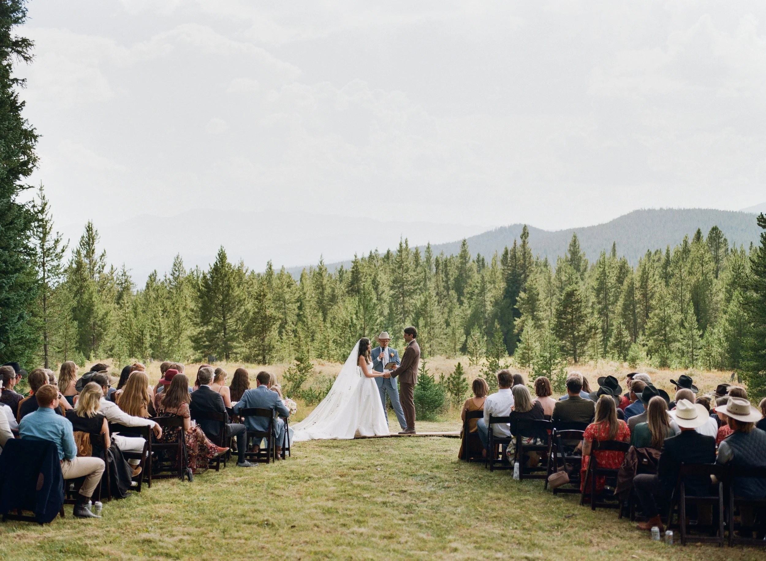 A wedding ceremony taking place outdoors in a forested area with mountains in the background. The bride and groom are standing in front of a small crowd, with an officiant between them. Guests are seated in rows of chairs, many wearing hats, with the scene set against a partly cloudy sky.