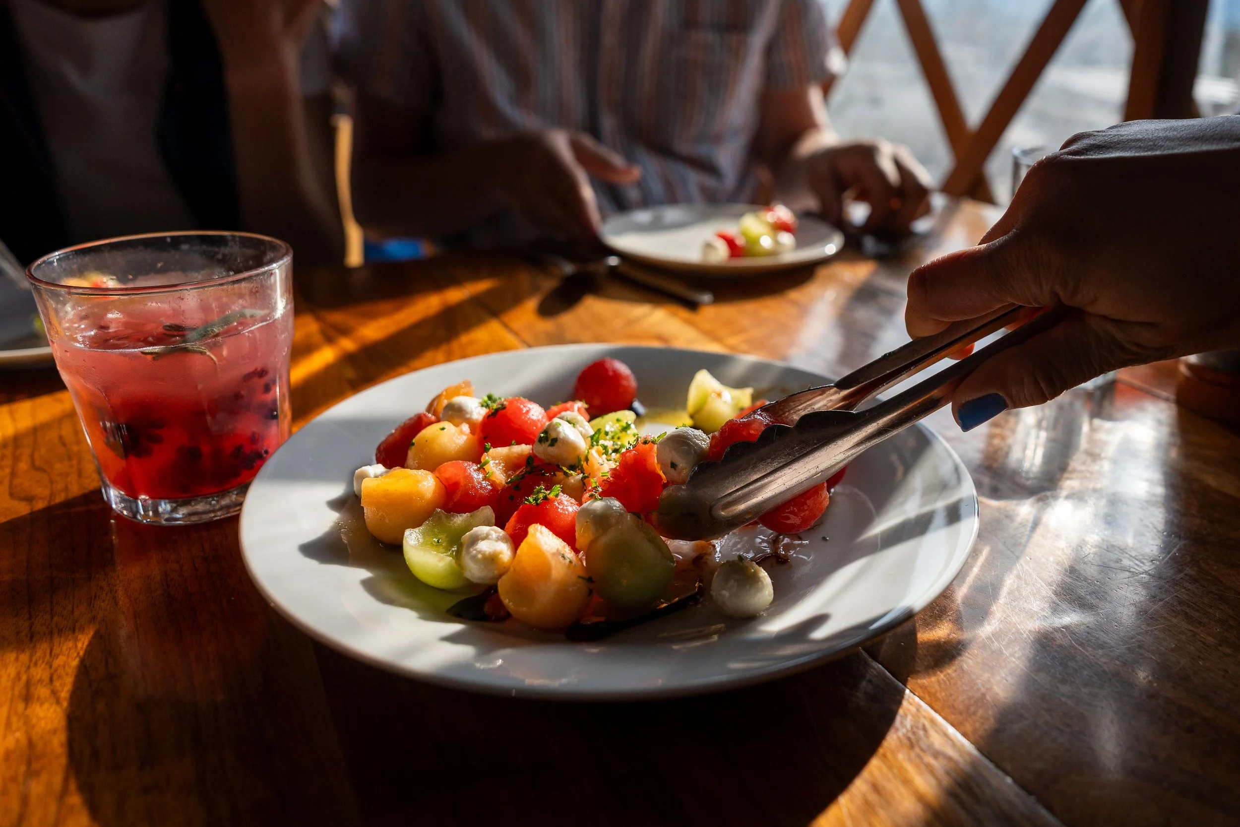A plate of mixed melon balls, cherry tomatoes, and mozzarella cheese with fresh herbs, on a wooden table. A person's hand is reaching with tongs to pick up food from the plate. In the background, there is a glass of berry-flavored beverage with fruit and herbs inside, and two people sitting at the table near a window.