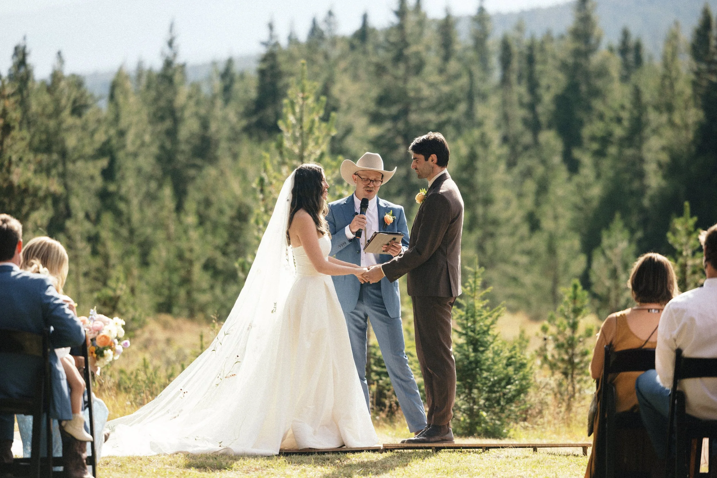 A couple getting married outdoors, holding hands and facing each other in front of an officiant. The bride is in a white gown with a veil, and the groom is in a brown suit. The officiant is wearing a cowboy hat and glasses, holding a microphone and a book. Guests are seated and watching the ceremony with a forest and mountain background.