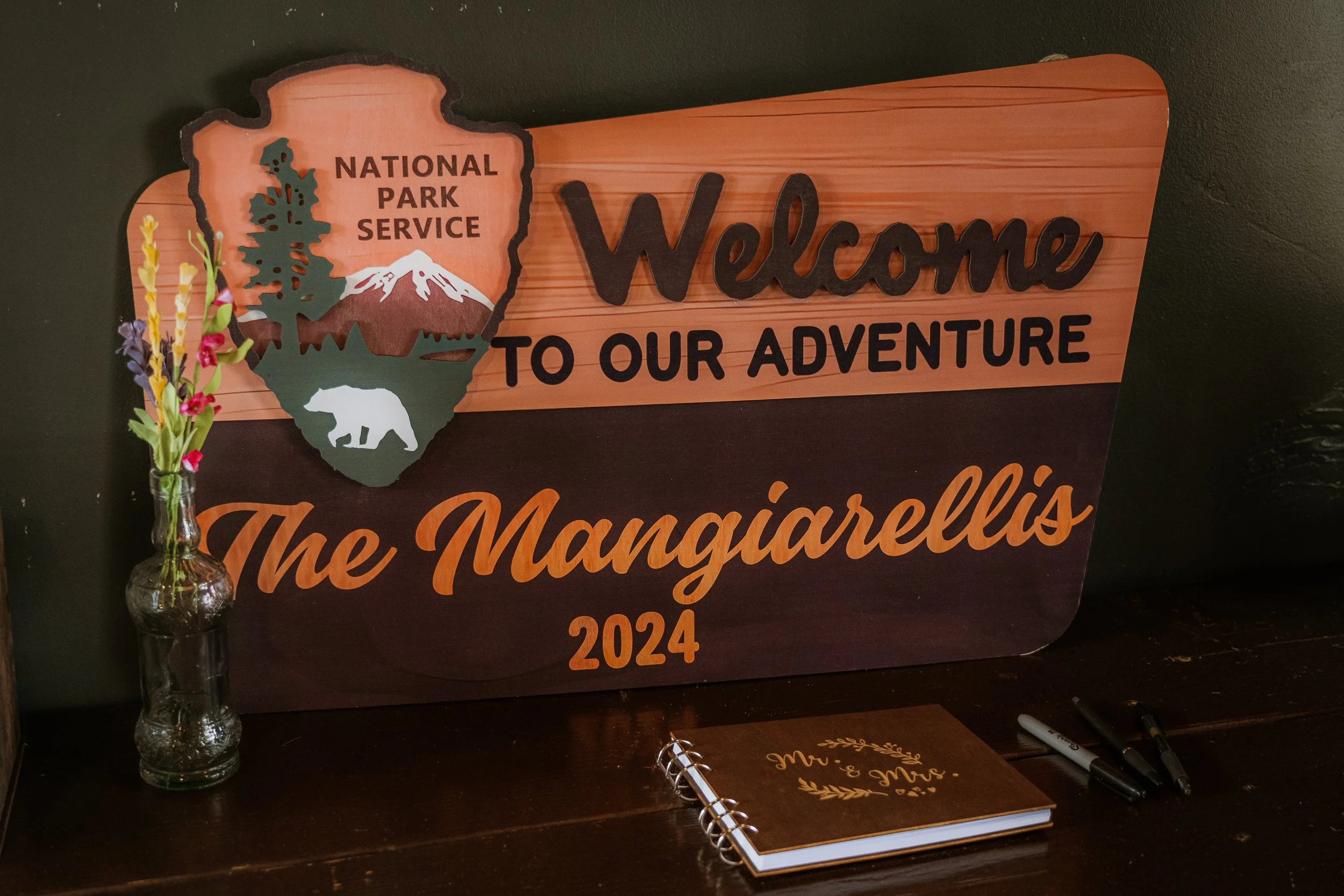 Wooden sign with the words 'Welcome to our adventure' and the National Park Service logo featuring a tree, mountain, and bear, on a black background.