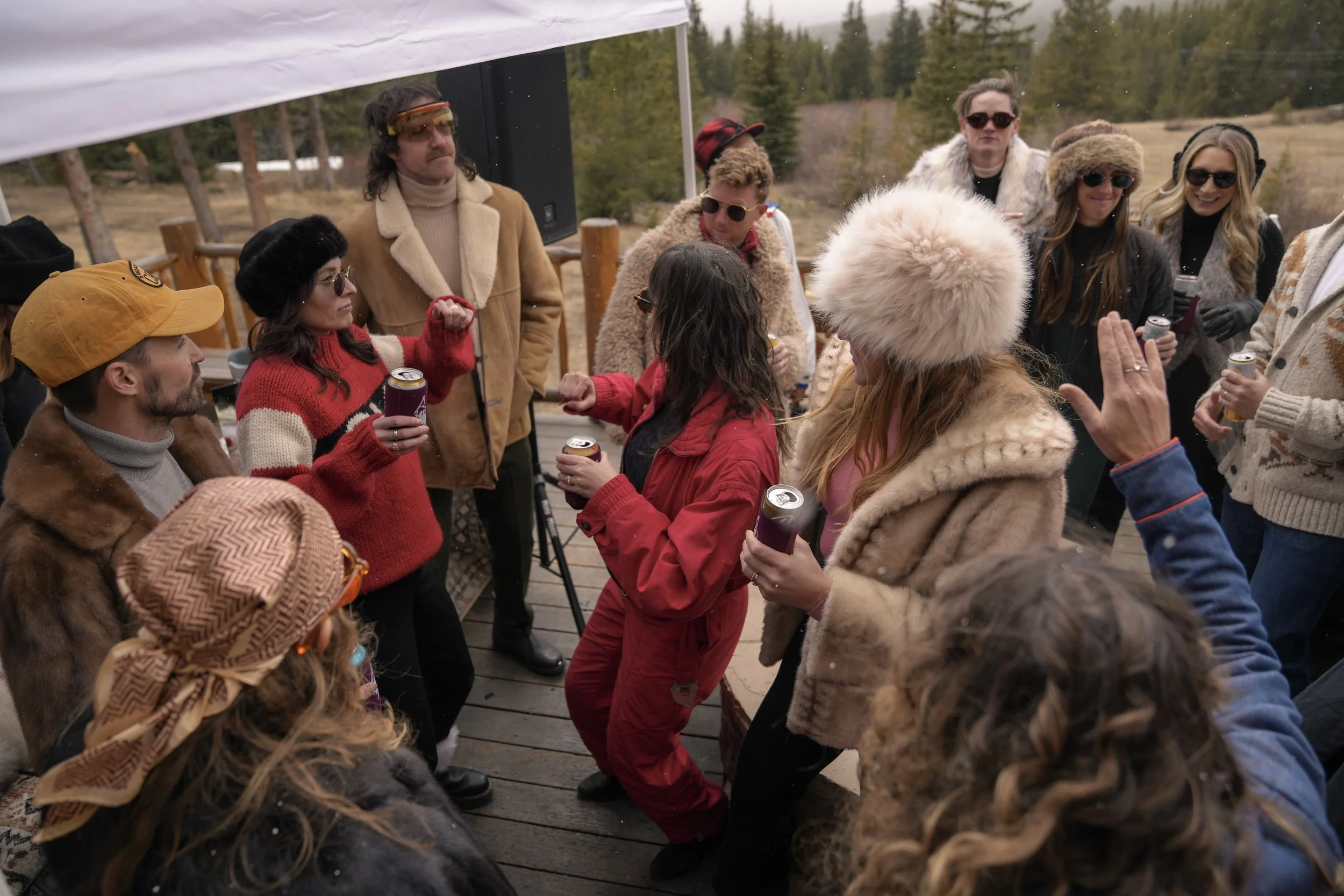 A group of people dancing and socializing outdoors near a wooden deck, surrounded by trees and mountains, during a chilly day.